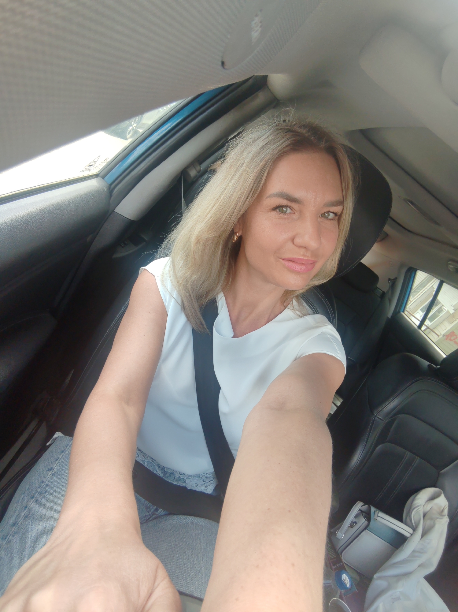A woman with blonde hair is sitting in the driver's seat of a car, taking a selfie. She is wearing a white t-shirt and jeans, and her seatbelt is fastened. The car appears to be parked, as indicated by the presence of buildings visible through the passenger window. The interior of the car is visible, including the dashboard and seats. The woman is smiling and looking at the camera. There is no information available about the specific location of Petropavlovsk-Kamchatsky, Russia in the image.