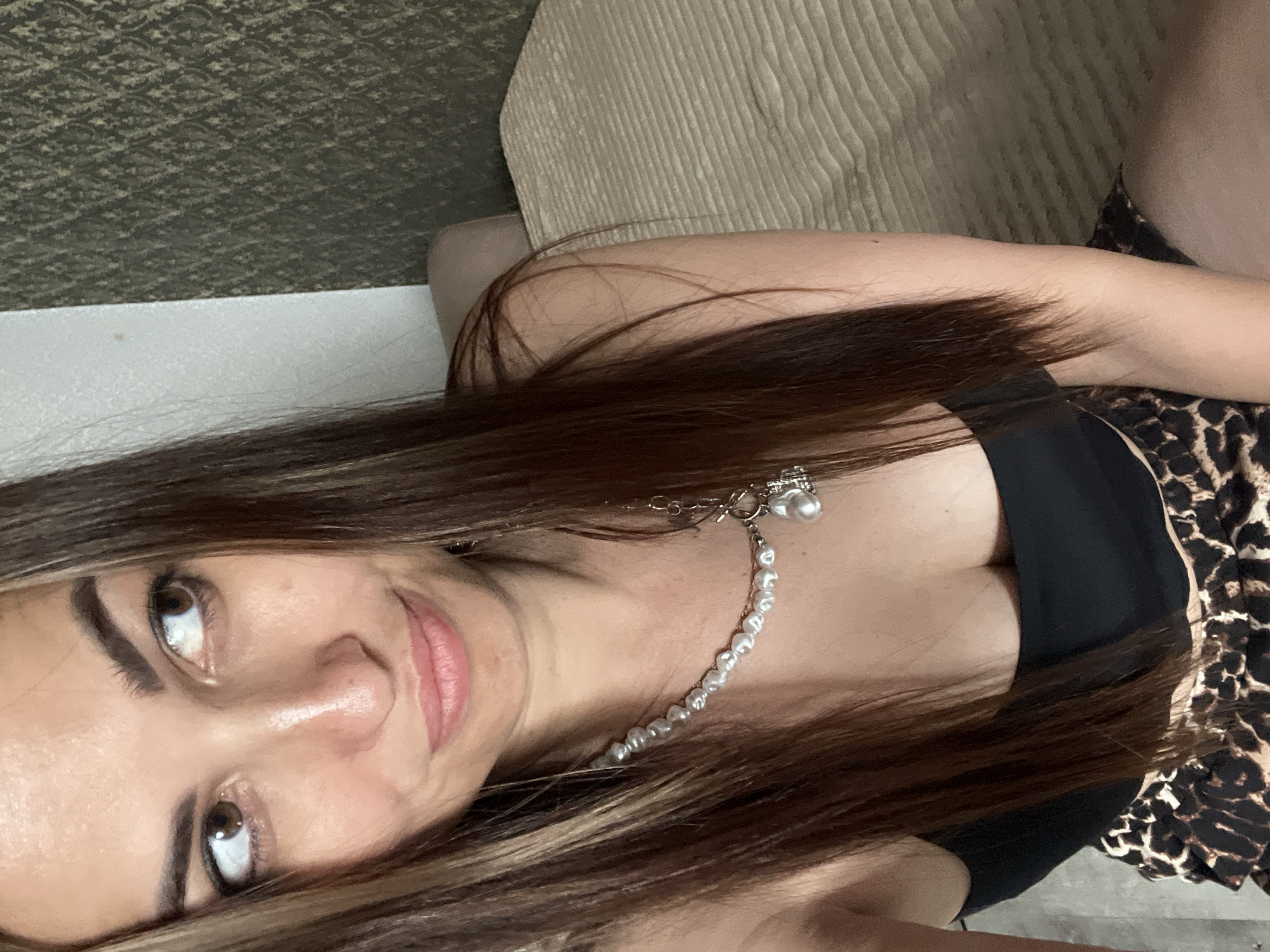 A woman with long, dark brown hair and brown eyes is pictured from the chest up, indoors. She is wearing a black top and a pearl necklace with a pendant. Her gaze is directed to the left of the frame, and she has a slight smile. The background appears to be a wall with textured wallpaper and a bed with a light-colored, textured spread.