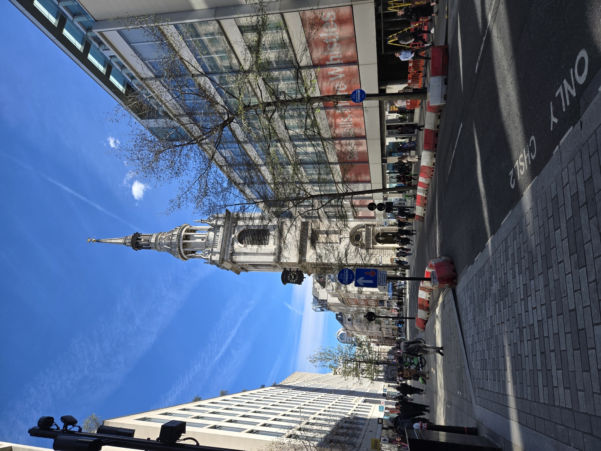 This image presents a detailed view of Cheapside, located in the City of London, United Kingdom, observed from a low angle on the northern sidewalk facing eastward. The scene is illuminated by strong sunlight under a clear blue sky featuring thin contrails and minimal cloud cover.
Dominating the central mid-ground is St. Mary-le-Bow church, a historical light-colored stone structure with a tall, ornate spire capped by a golden weather vane. A dark clock face is visible on its tower. Bare deciduous trees are positioned in front of and adjacent to the church.
To the south (right) of the church, a modern, multi-story building with a facade of large glass windows and light grey panels is prominent. A large red banner, partially displaying white text including "Whistles," covers a section of its lower floors. Beneath this banner, several ground-level retail units are visible, including "Greggs." Orange and white plastic road barriers are arrayed along the curb in front of this building, and a yellow ladder suggests ongoing maintenance or construction work.
To the north (left) of the church, a tall, light-colored building with numerous rectangular windows extends down the street. A ground-floor storefront with "itsu" branding is visible. The northern sidewalk in the foreground is paved with grey rectangular bricks.
The asphalt roadway in the foreground and mid-ground is marked with white lines, including a partially visible inverted phrase "ONLY CHISLE 2" (likely "CHISWELL ONLY") indicating a specific lane designation. Additional orange and white plastic barriers separate sections of the road. Street signage visible includes a blue circular sign denoting a bus and cycle lane, and a blue rectangular sign with an upward arrow and the text "Priority over oncoming vehicles."
Approximately twenty pedestrians are visible on both sidewalks, walking in various directions. One individual, a male in a light grey top and dark trousers, is walking towards the camera on the northern sidewalk. Strong, elongated shadows are cast across the street and sidewalks by the buildings and trees, indicating bright sunlight originating from the south. The scene depicts a busy urban commercial environment with a juxtaposition of historical and contemporary architecture.