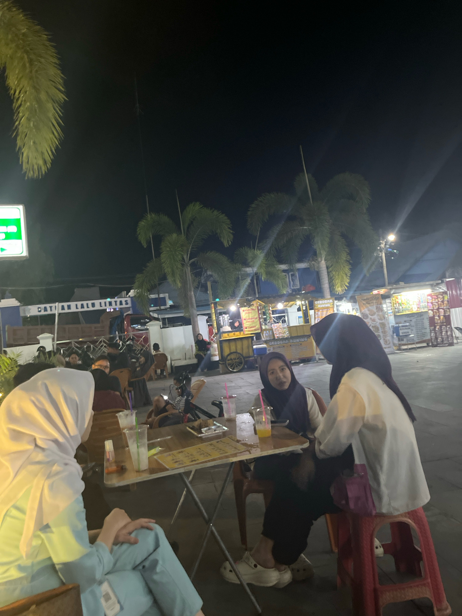 The image depicts an outdoor night scene in Sengkang, Indonesia. In the foreground, three individuals are seated at a table, engaged in conversation and consuming beverages. One person, wearing a white hijab and light blue attire, is partially visible from the side. Another individual, in a dark hijab and white shirt, is facing the viewer. The third person, also in a dark hijab, is seated opposite them. On the table, there are glasses with drinks, a plate of food, and a menu. The background features several food stalls with lights, palm trees, and a building with signage that appears to read "PUSAT LALU LINTAS." The atmosphere suggests a casual gathering at an eatery or night market.