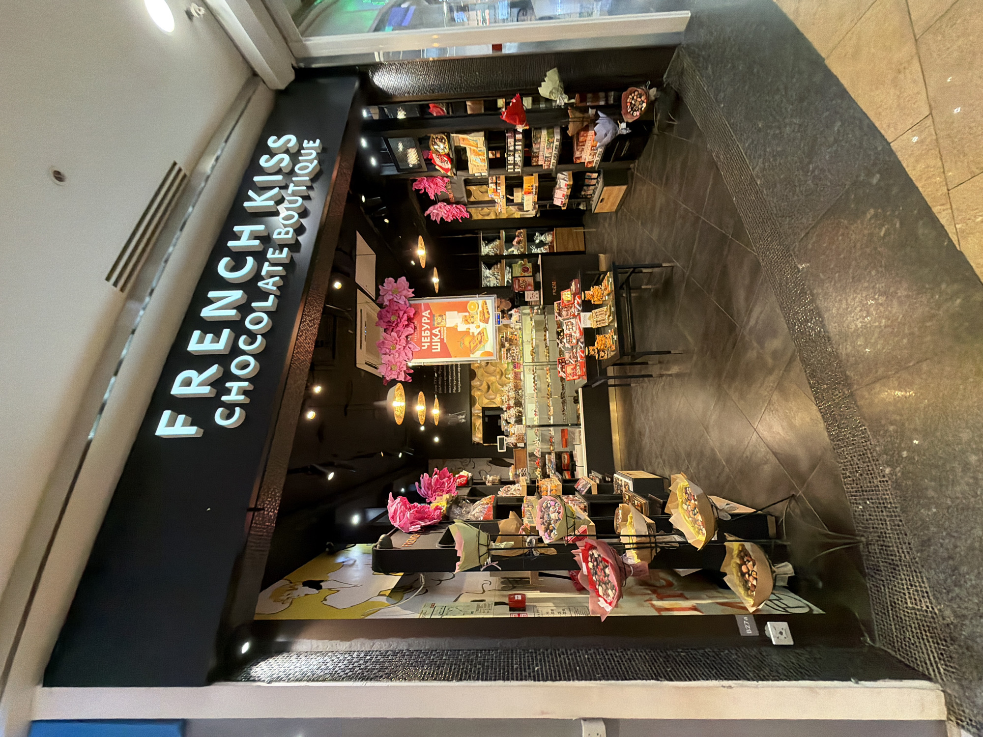 The image shows a tilted shot of the storefront of "FRENCH KISS CHOCOLATE BOUTIQUE." The location appears to be indoors, possibly within a mall or shopping center. The store's signage is prominently displayed in white lettering. Inside, shelves are stocked with various products, seemingly chocolates and related items. Decorations include pink floral arrangements. The storefront features a patterned facade. The overall setting seems to be a commercial retail space. A sign in the shop reads "ЧЕБУРАШКА" which suggests this is in Moscow, Russia.