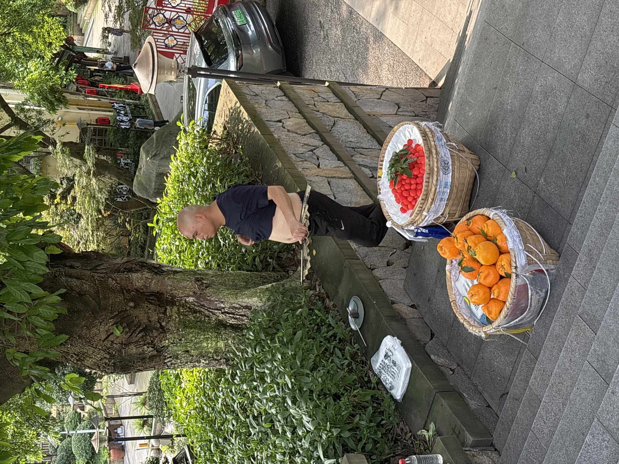 In Yuzhong District, China, an individual stands between two baskets of fruit. The upper basket contains small, red fruits, while the lower basket is filled with oranges. The person is cutting fruit with a knife. A small dish with cut fruit and a spoon are located near the person. A plastic bag is also on the ground near the fruit. The scene is outdoors, adjacent to a tree, and includes concrete steps, foliage, and a stone wall. A street and parked car are visible in the background.
