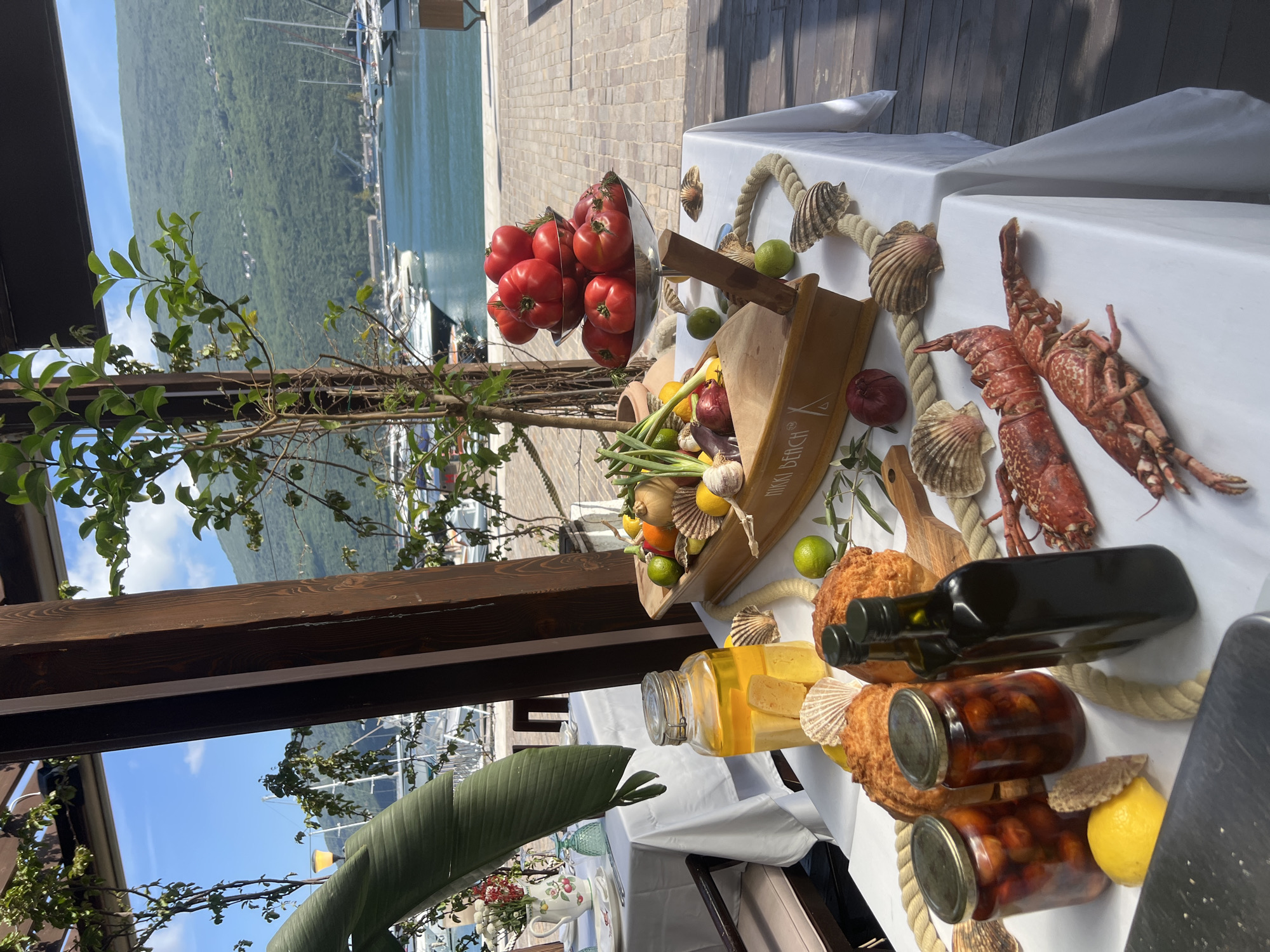 This image captures a vibrant outdoor scene at a waterfront location in Đenovići, Montenegro, during bright daylight hours. The main subject is a meticulously arranged table, covered with a white tablecloth, laden with fresh ingredients and food items.
On the table, a large glass bowl is filled with ripe red tomatoes. Two cooked lobsters are prominently displayed, alongside decorative nautical ropes and various seashells, suggesting a seafood theme. A unique wooden boat-shaped container holds an assortment of fresh produce including onions, garlic, lemons, limes, and spring onions. A large glass jar contains a yellow liquid, possibly oil, with what appear to be cheese cubes, and two smaller glass jars filled with preserved items like olives or cherry tomatoes are also present. A dark bottle, likely olive oil, stands near some round, golden-brown pastries or breads.
The background reveals a picturesque coastal landscape: a marina with numerous boats and yachts on calm blue water, framed by lush green mountains under a clear blue sky with scattered white clouds. To the left, green foliage from plants and a dark wooden pergola structure are visible, contributing to the outdoor setting. The ground appears to be paved with light-colored stone or tile.
The arrangement suggests a fresh and abundant culinary presentation, likely a display of local ingredients for a meal or a special event. The visible text "NIKKI BEACH" is inscribed on the side of the wooden boat container. No people are visible in the image. The overall atmosphere is inviting and highlights natural beauty and fresh produce.