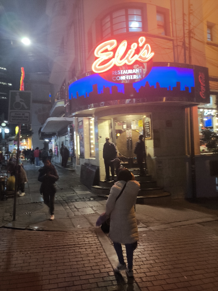 A street scene at night. The main focus is on "Eli's Restaurant Confiteria" with its bright neon sign. People are walking on the sidewalk in front of the restaurant. A person in a white coat stands facing away from the camera on the brick sidewalk. To the left, a person in a dark coat walks towards the restaurant while looking at their phone. Other pedestrians can be seen further down the street. A handicap sign is visible on the left.