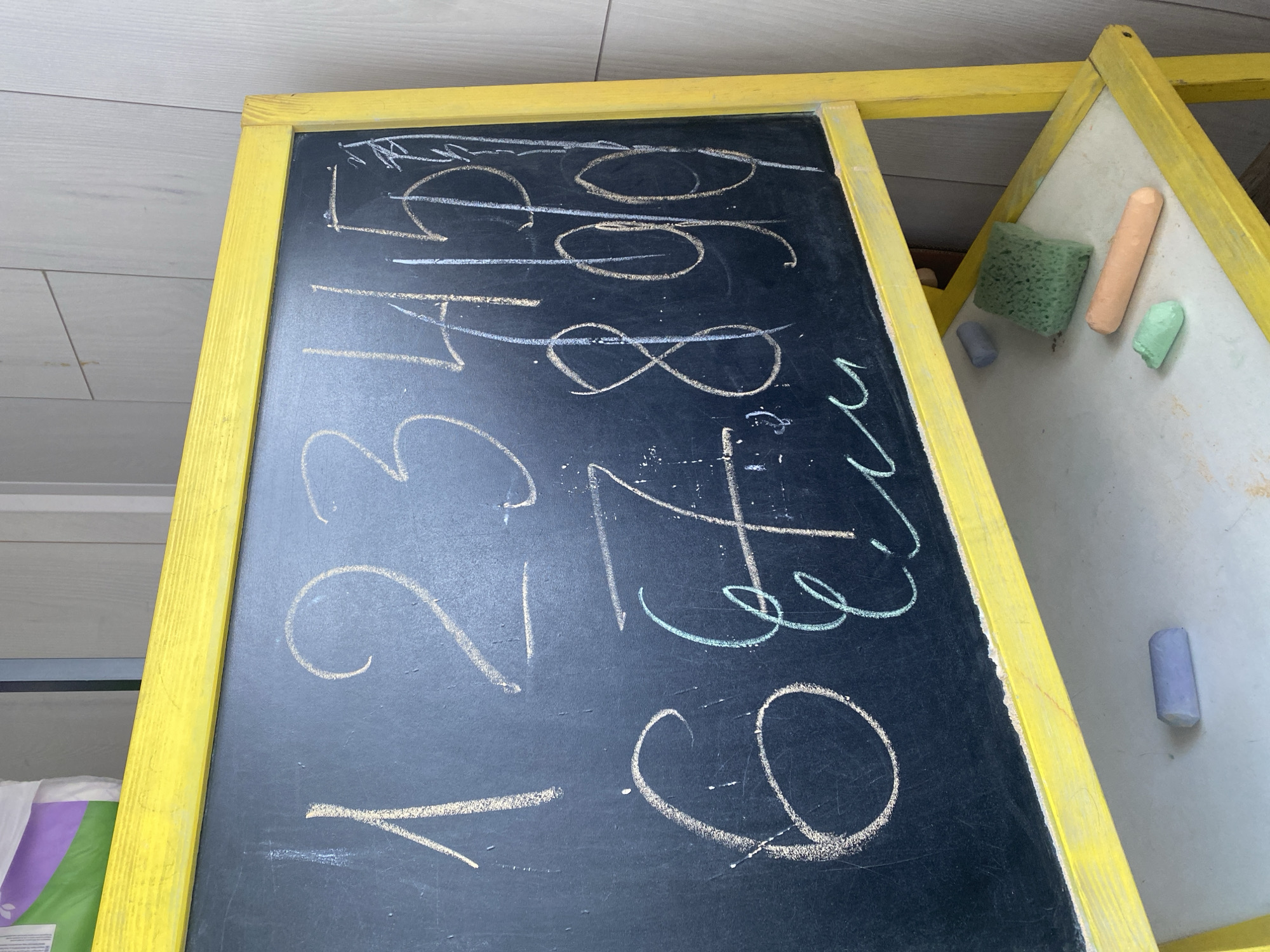 This is a medium shot, close-up, eye-level photograph of a child's chalkboard easel with chalk drawings. The chalkboard is framed in bright yellow wood. On the black chalkboard, several numbers are drawn in white chalk, including a large "1", "2", "3", and "4". There are also some circular shapes drawn in yellow and white chalk, some of which appear to be numbers such as "0", "6", and "8". There is also a drawing of an infinity symbol in white chalk and what looks like a "4" or "F" in light blue chalk. To the right of the chalkboard, part of a dry-erase board is visible, also framed in yellow wood. On this board, there are several pieces of chalk and a green sponge. The chalk pieces are orange, light green, and purple.
The scene appears to be indoors, possibly in a play area or classroom, given the presence of the chalkboard and dry-erase board. The background is blurred but seems to show a wall with horizontal wood paneling. There are no people visible in the frame. The lighting suggests it is daytime. The overall impression is one of a creative or educational space. The chalk drawings are somewhat messy, indicating they were likely drawn by a child.