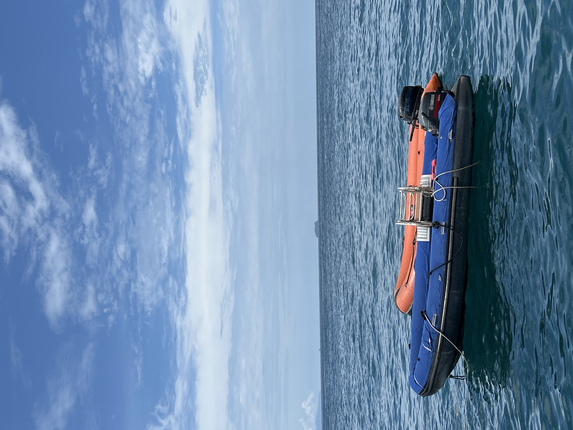 Two inflatable boats are floating on the water in Ao Nang, Thailand. The boat in the foreground is blue with a black inflatable tube and is partially covered with a blue tarp. A white rope is tied to the bow of the blue boat and extends to the left. A Yamaha outboard motor is attached to the stern of the blue boat. The second boat, partially visible behind the blue boat, is orange and also appears to be an inflatable boat with an outboard motor. The water is choppy and a distant landmass is visible on the horizon under a partly cloudy blue sky.