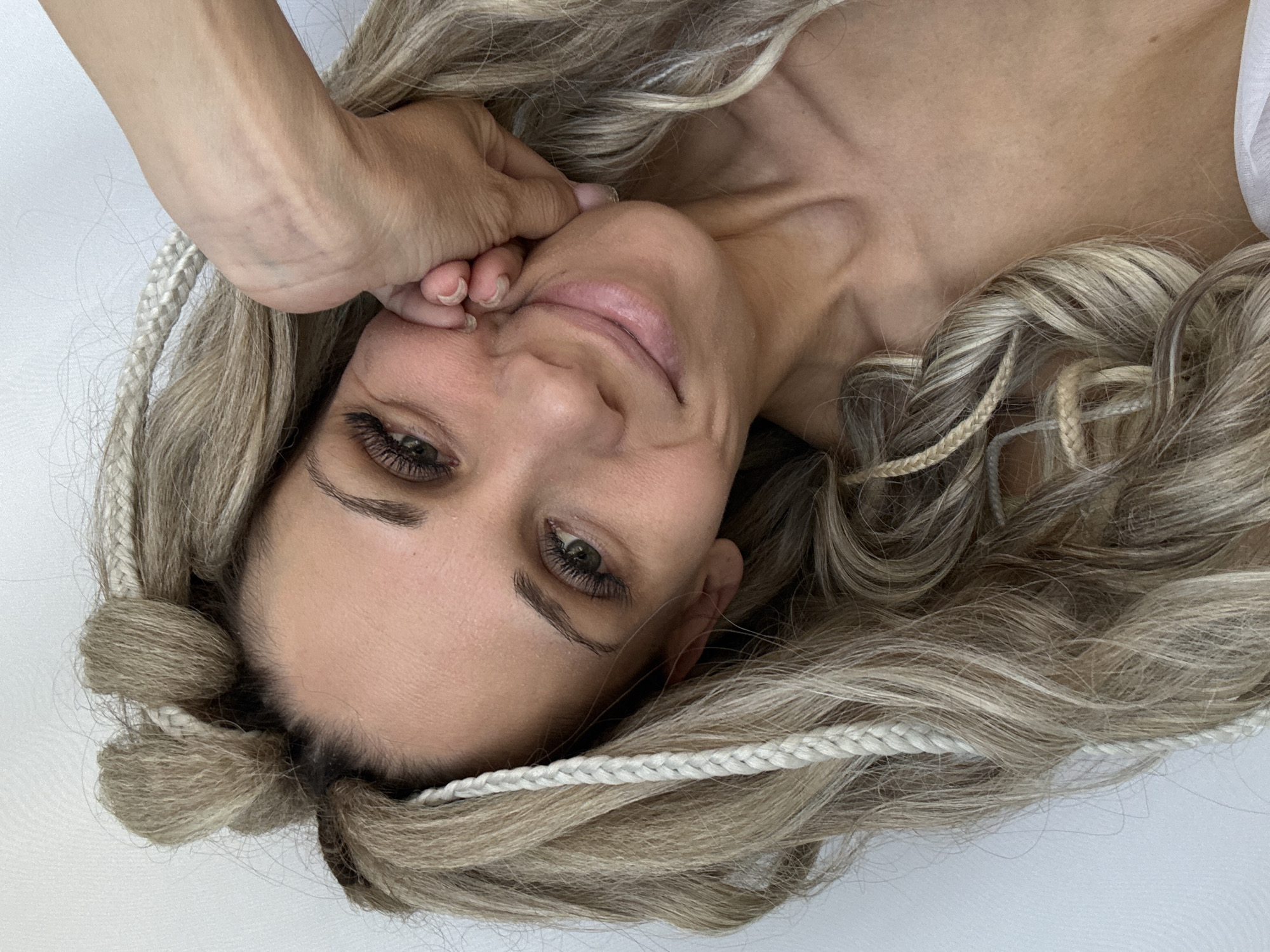 A woman is lying down with her head tilted to the right. She has blonde hair with white braids woven throughout. Her right hand is touching her face near her cheek. She is smiling softly. The background is white. It is not possible to determine the location in Rusanovo, Russia, based solely on the provided image.
