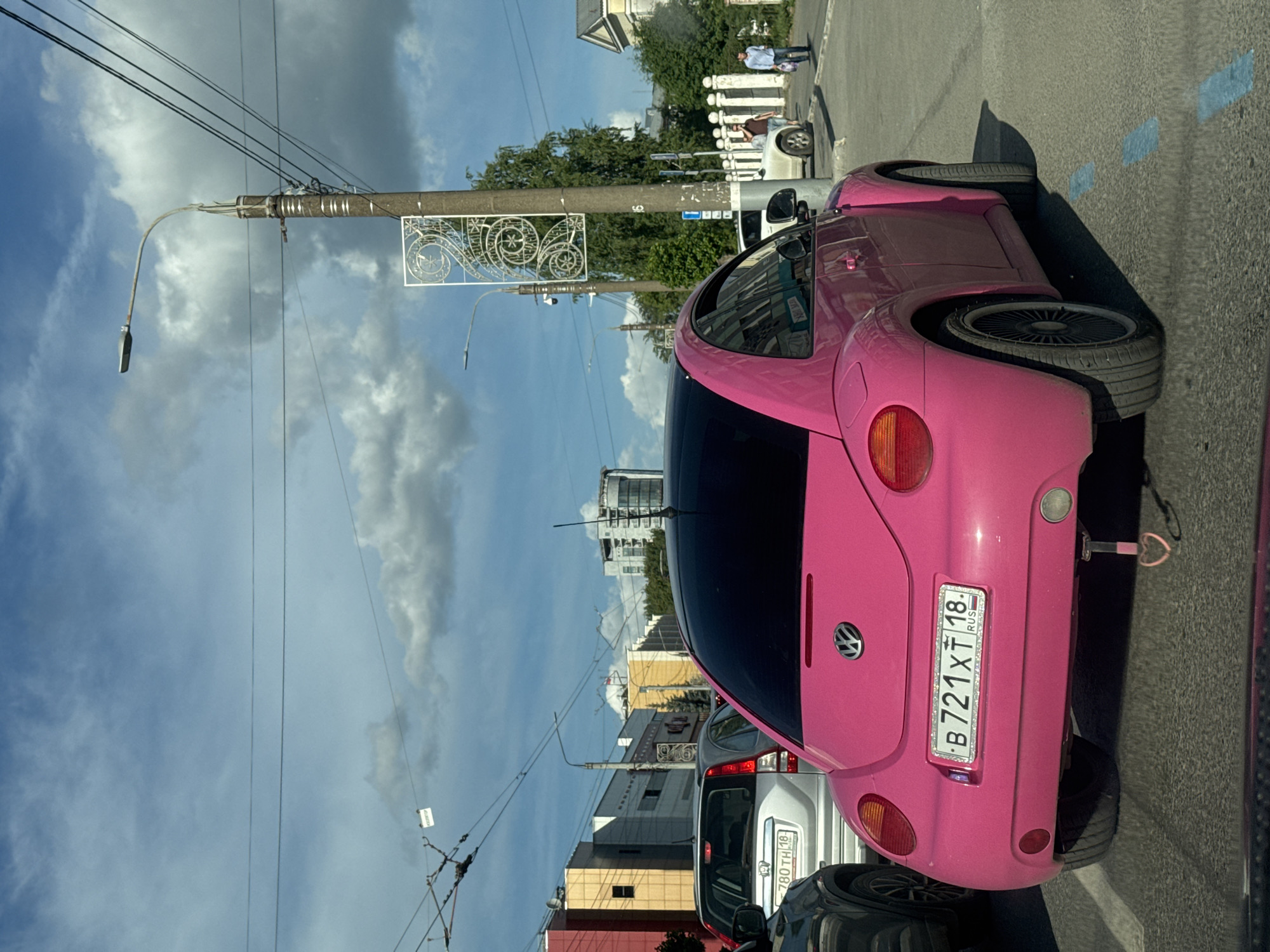 This daytime photograph, likely taken on a sunny or partly cloudy day, captures a street scene in Izhevsk, Russia. The primary subject is a vibrant pink Volkswagen Beetle, viewed from its rear-right side, appearing to be either parked or in traffic. This car features customized wide fenders, low-profile tires, and a blacked-out rear window. A small pink heart-shaped ornament hangs from its tow hitch.
Visible on the rear of the pink Beetle is a Russian license plate displaying "B 721 XT 18 RUS", with "18" indicating the Udmurt Republic (where Izhevsk is located). A white SUV or minivan is visible directly behind the pink Beetle, also displaying a partially visible Russian license plate: "780 TH 18 RUS". Part of a dark-colored car is seen in the foreground, in front of the pink Beetle.
The scene includes multiple concrete utility poles, some fitted with streetlights and decorative white metal panels featuring intricate swirling designs. Overhead, numerous power lines stretch across the sky. In the background, various buildings of different heights and architectural styles line the street, interspersed with trees and greenery. People are visible on the sidewalk to the right, with one person standing near a white car and another further in the distance. The road surface is asphalt with faded blue markings. The sky is a mix of clear blue patches and white, fluffy clouds.