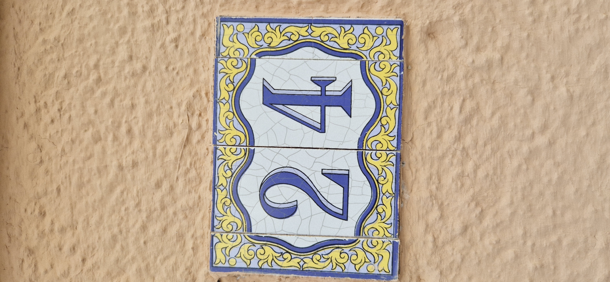 A close-up, eye-level, vertical outdoor shot shows a decorative house number sign mounted on a textured, peach-colored stucco wall. The sign is composed of two rectangular ceramic tiles, side-by-side. Each tile has a blue and yellow ornamental border with a repeating scroll pattern on a white background. The left tile displays a large blue numeral "2", and the right tile displays a large blue numeral "4". The numbers are also outlined in a darker blue. The stucco wall has a rough, uneven texture with small indentations and visible cracks. The lighting is neutral, suggesting it is daytime, and there are no people or other objects visible in the frame.