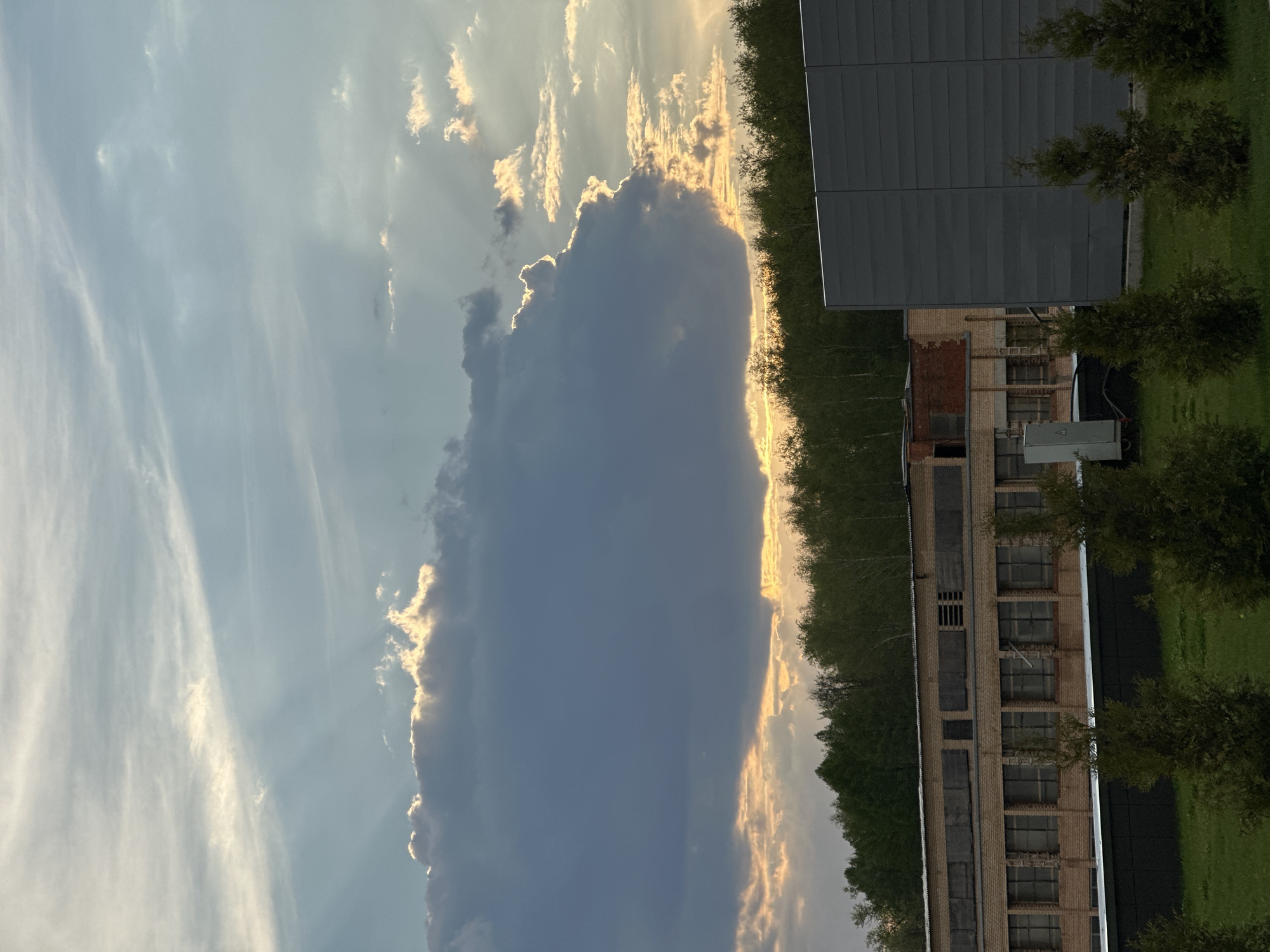 The image presents a scene in a place with a large cloud formation covering the sky, with a dark grey cloud in the foreground contrasted against a sky of light blue and white. The sun is visible, and there are sunbeams breaking through some of the clouds.
On the right side of the image, there is a building with a grey roof and a brick facade, featuring boarded windows. A line of trees separates the building from the cloud formation. There's also green grass visible, likely a lawn area.
Based solely on the image, it is not possible to confirm the location is Moscow, Russia, or provide any information about people, actions, or interactions.