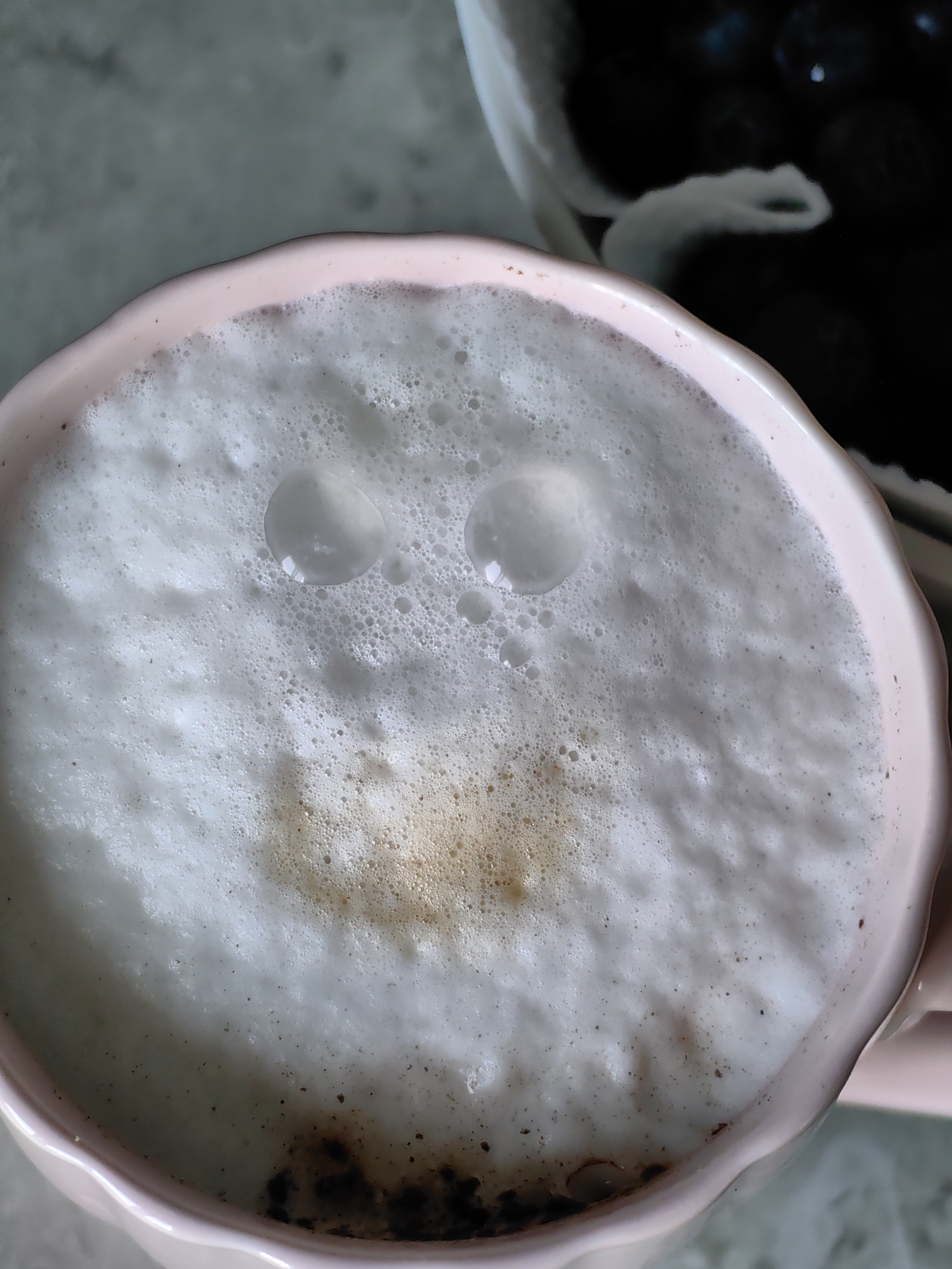 The image shows a close-up, overhead view of a pink mug filled with frothy white coffee. Two large bubbles on the surface of the foam resemble eyes, and a lighter patch of coffee beneath them suggests a smiling mouth, creating a face-like appearance. The mug has a decorative, scalloped rim. In the background, out of focus, is a dark object, possibly a container of blueberries or other dark berries, and a blurred surface that appears to be a countertop. The scene is indoors, with neutral lighting. There are no people visible in the image. This image was taken in Fanipal, Belarus.