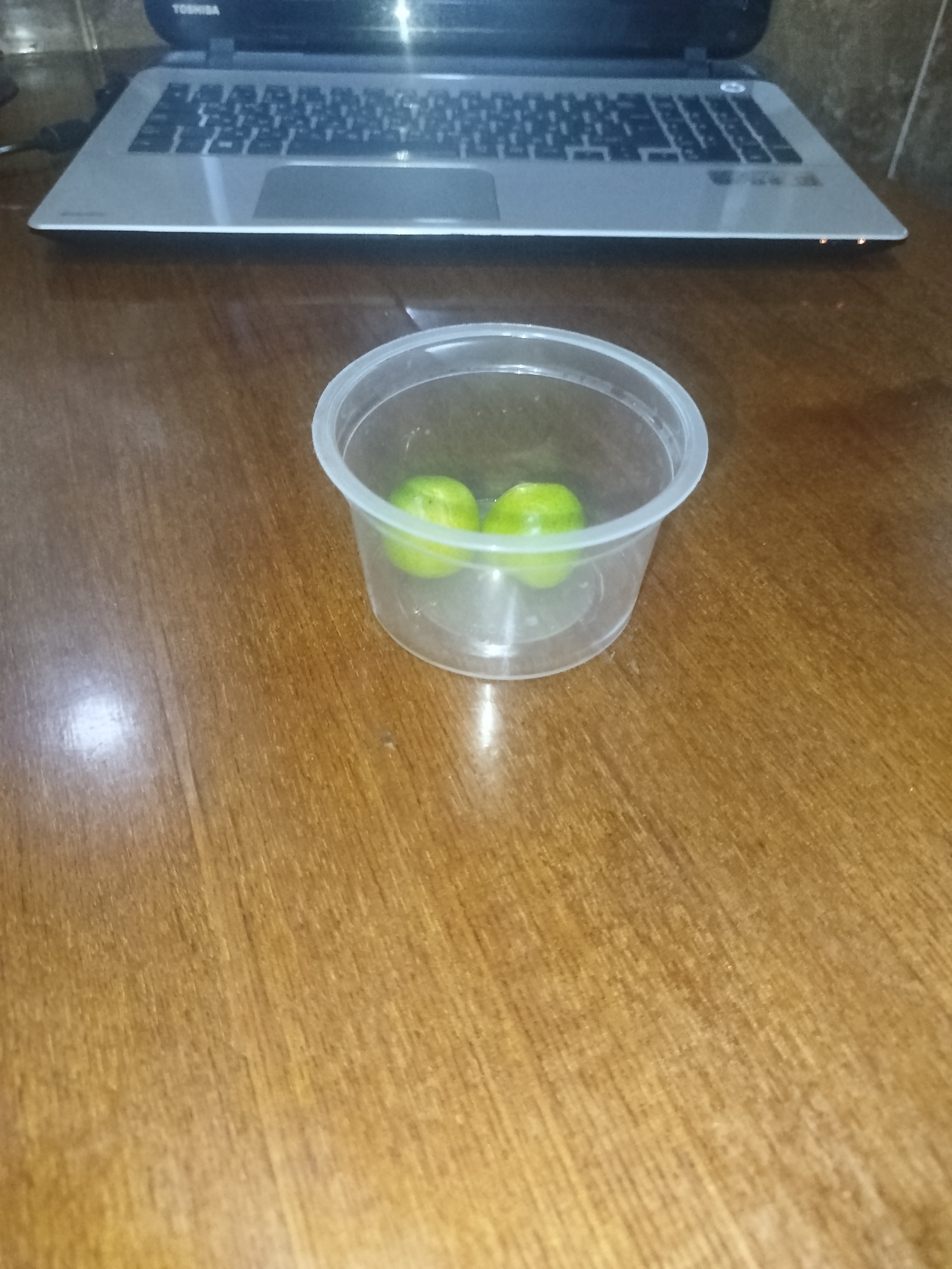 A clear plastic container holding two small green limes sits on a wooden surface. A laptop is visible in the background, positioned behind the container. The scene appears to be indoors, possibly on a desk or table. There is no indication of the location in the city of Sabunchu, Azerbaijan.