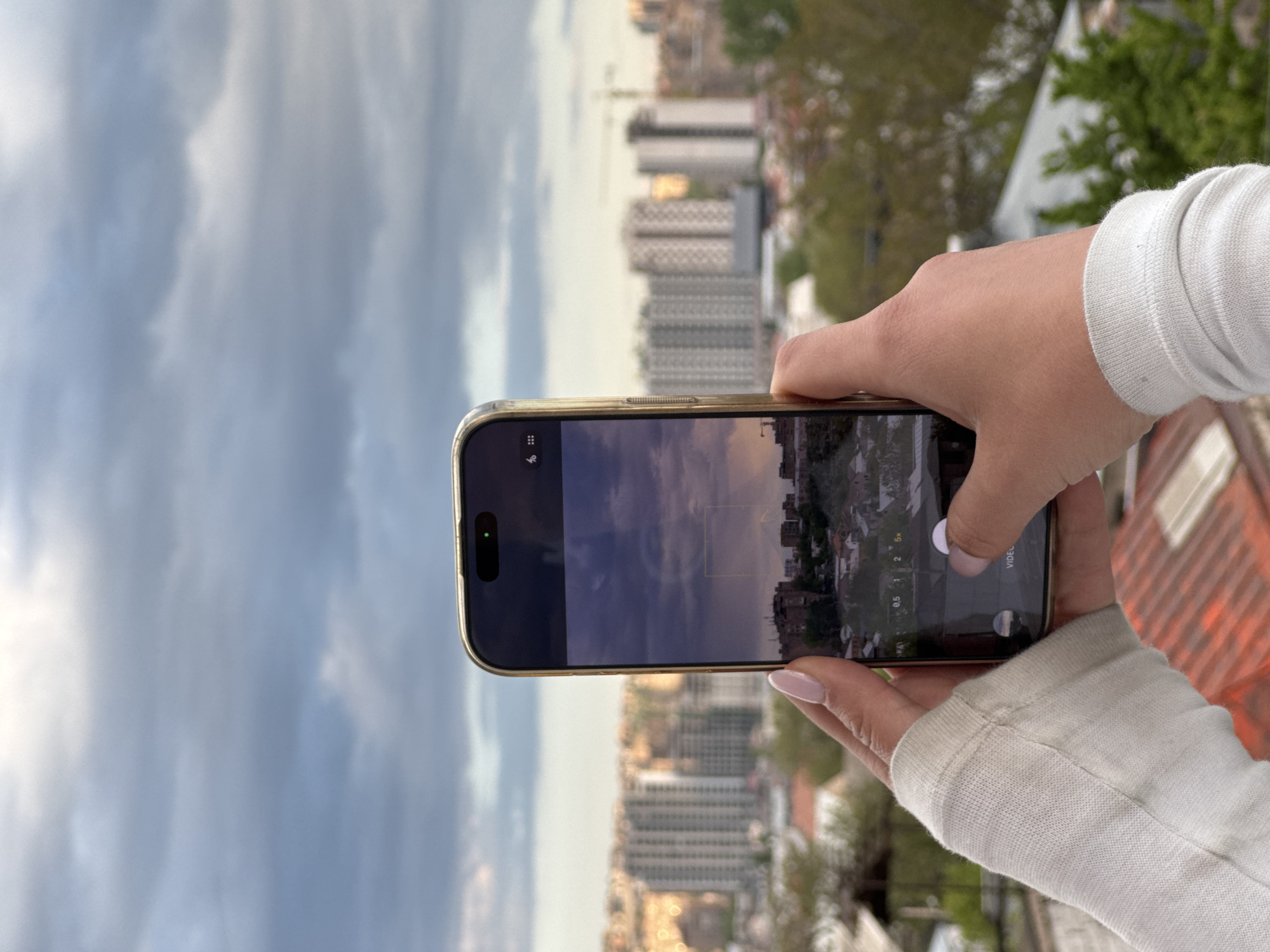 A person is holding a gold iPhone in their hands, taking a photograph of the urban landscape of Arinj, Armenia. The phone screen displays a dusk or dawn cityscape. The background beyond the phone features buildings of various heights, interspersed with trees, all under a cloudy sky. The angle of the shot suggests the person is on a higher vantage point, possibly a rooftop or elevated balcony. The person's hands are visible, with light pink nail polish. The left arm is wearing a light-colored sweatshirt with ribbed cuffs. The orange safety material is visible.