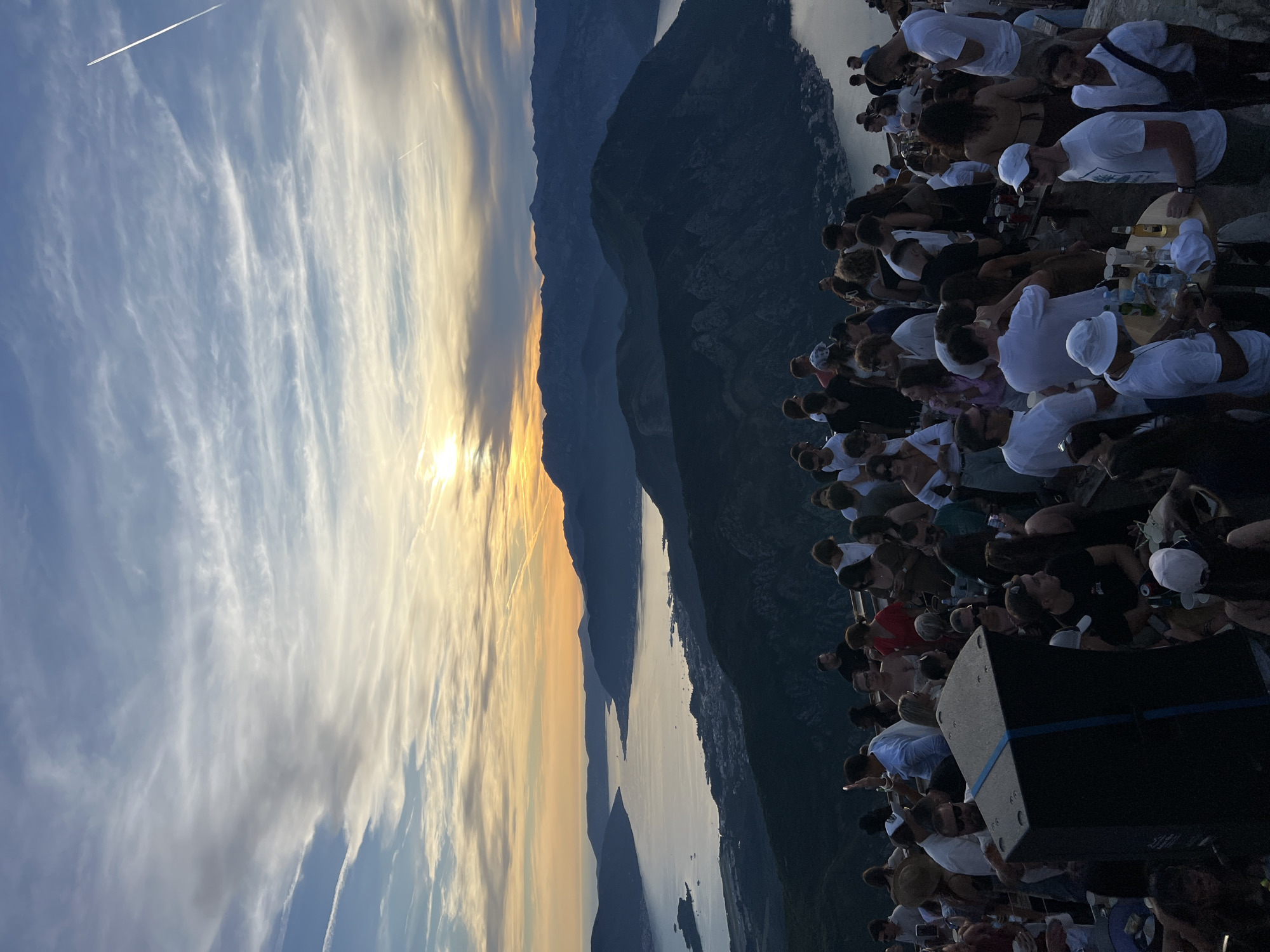 The image depicts an outdoor gathering of a large group of people at sunset, overlooking a bay with mountains in the background. The location is identified as a viewpoint near Žanjev Do, Montenegro. The sun is low on the horizon, casting a warm orange and yellow glow across the sky, which is filled with wispy clouds and contrails from airplanes.
The foreground is occupied by a crowd of people, many of whom are dressed in casual attire. They are standing and mingling, with some holding drinks and others taking photographs. Several tables are visible, laden with bottles and cups, suggesting a social event or party. A large black speaker box is positioned in the mid-ground, with blue tape around it. The people are diverse in age and gender, and their interactions appear relaxed and social.
The mid-ground shows the dramatic landscape of dark, rugged mountains sloping down to a wide expanse of water, likely a bay or fjord. The water reflects the colors of the sunset. A coastal town or settlement is visible along the edge of the water. The overall atmosphere is festive and scenic, capitalizing on the stunning natural setting and the beautiful sunset.