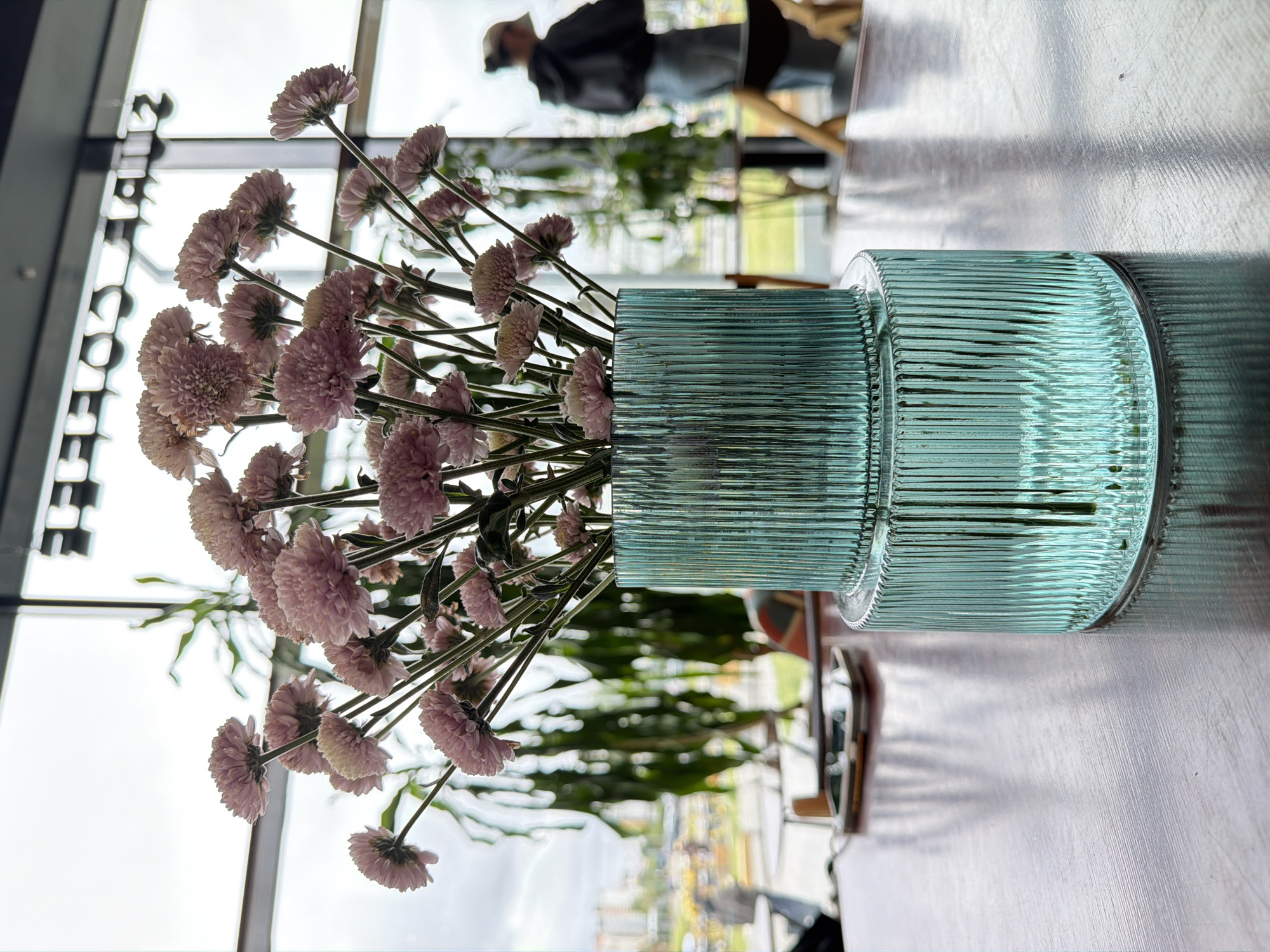Here's a description of the image:
The image shows a bouquet of pink chrysanthemums in a ribbed, aqua-colored glass vase. The vase is sitting on a light-colored, possibly metal, table. The background is blurred but suggests a bright interior space, possibly a cafe or restaurant, with large windows overlooking an urban setting. A person is visible through the window, wearing dark clothing and a hat, and appears to be walking outside. Reflections are visible in the glass surfaces of the window. The scene is well-lit, with natural light coming from the windows.
There is no information that would lead me to believe this is Moscow, Russia.