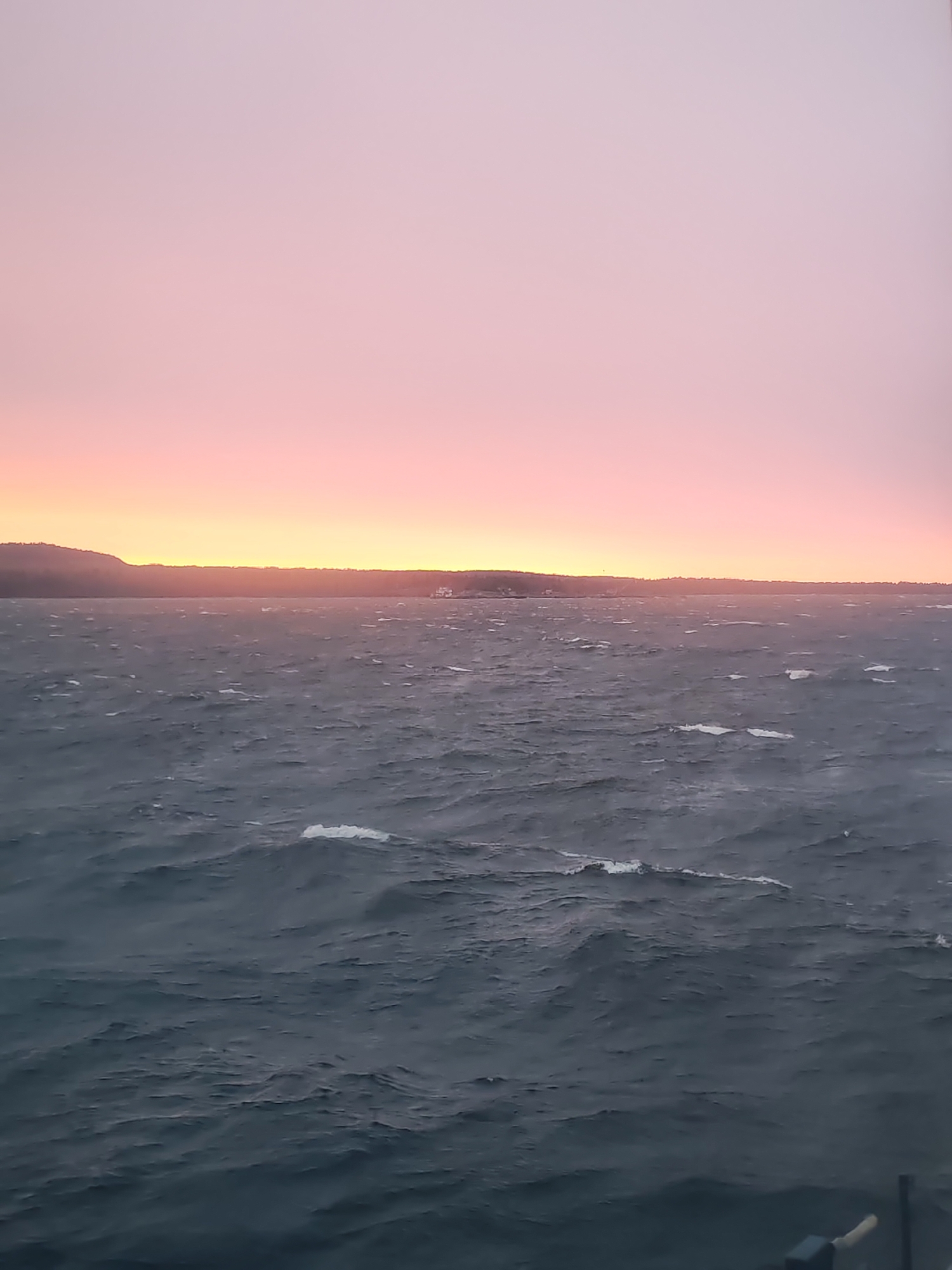 The image displays a panoramic view of a vast body of water, likely a lake or sea, under a striking sky during either sunrise or sunset. The sky dominates the upper two-thirds of the frame, showcasing a beautiful gradient of colors, transitioning from a soft, light pinkish-purple at the top to vibrant orange and yellow tones along the horizon.
A dark, undulating landmass, possibly a coastline with hills or forests, stretches across the distant horizon, forming a stark silhouette against the brightly colored sky. A small, indistinct white structure, potentially a building or lighthouse, is faintly visible on this distant landmass towards the right of the center.
The lower third of the image is filled with the expansive water, which appears dark blue-grey and very choppy. Numerous white caps and streaks of foam are visible across the water's surface, indicating strong winds and rough conditions. In the bottom right corner, a dark, out-of-focus object, possibly a railing or part of a boat structure, is partially visible.
No people are present in the scene, and no visible text can be identified. The overall impression is one of powerful natural beauty, with the dramatic, colorful sky contrasting with the wild, turbulent water. Given the location context of Shcheleiki, Russia, this view could be of a large body of water such as Lake Ladoga.