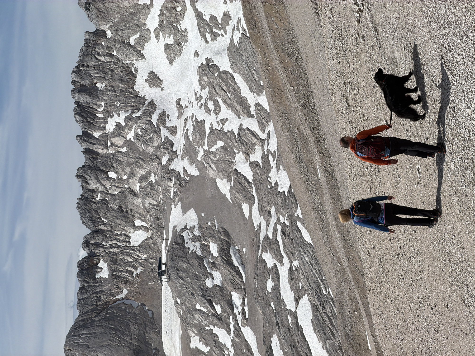 In Wildermieming, Austria, two individuals, a woman and a man, walk with a black dog on a gravel path. The woman, wearing a blue jacket and a backpack, is on the left. The man, in an orange jacket and also carrying a backpack, is to her right, holding a leash attached to the dog. They are walking away from the camera, descending a slope. The background features a stark, rocky mountain range with patches of snow, and a clear sky above. A ski lift station is visible on a higher ledge in the mid-ground. The terrain is rugged and appears to be at a high altitude, suggesting a mountainous environment suitable for hiking or skiing.