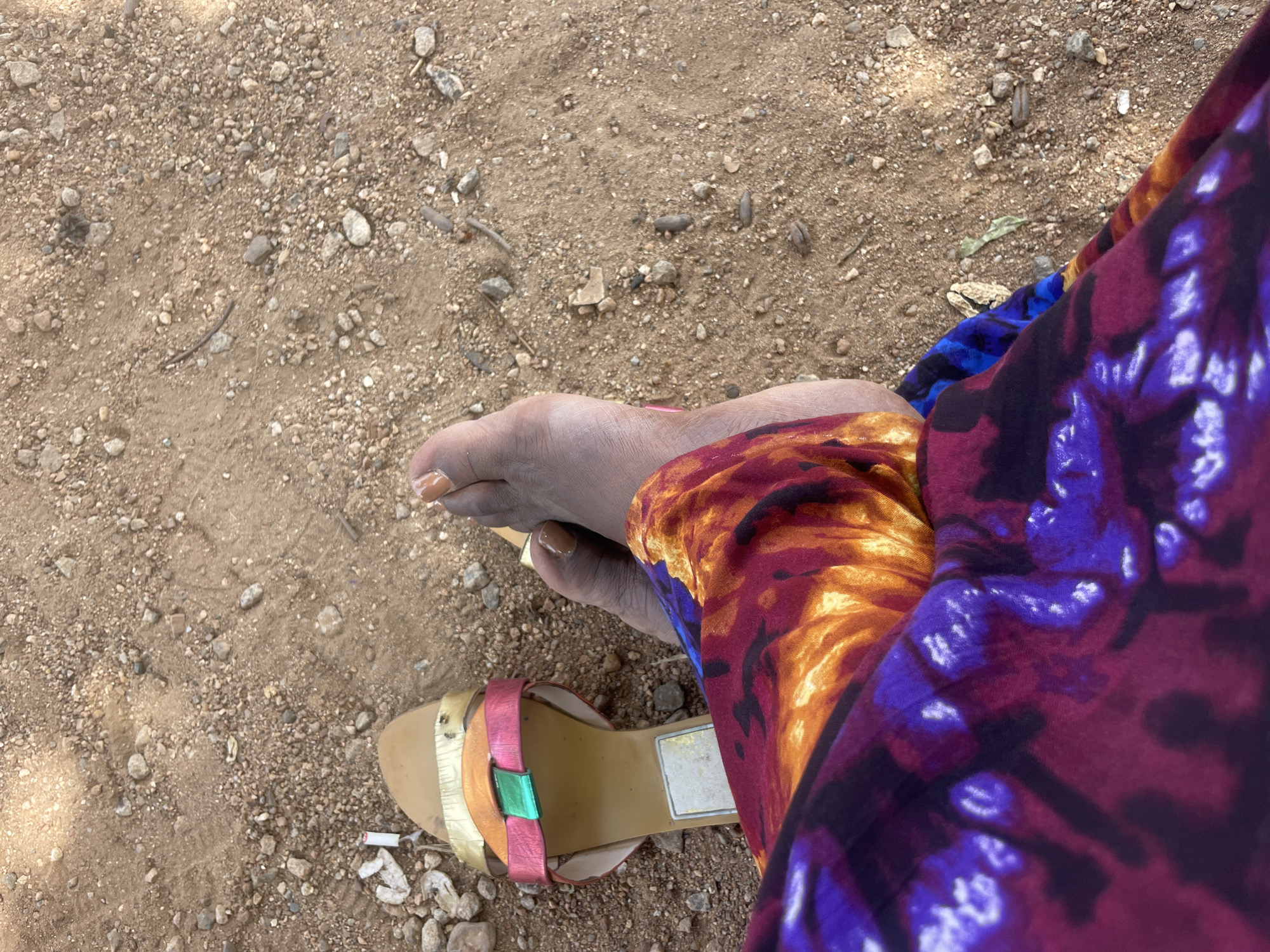 The image shows a person's legs and feet resting on a sandy and rocky ground. The person is wearing a colorful sandal with gold, pink, and green straps on their right foot, and their left foot is bare with painted toenails. They are also wearing a vibrantly patterned garment with shades of purple, orange, and yellow. The ground appears to be a natural outdoor environment, possibly a pathway or unpaved area in Bauchi, Nigeria, given the dry, sandy soil and scattered stones. The lighting suggests it is daytime.