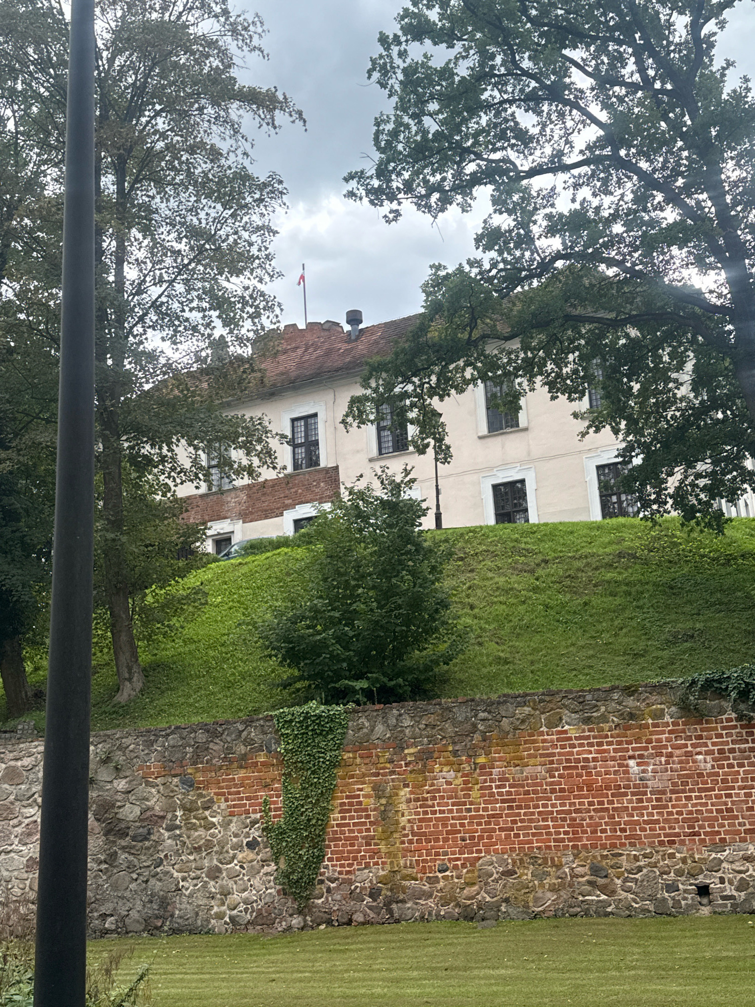 The image depicts the exterior of the Łagów Castle in Łagów, Poland. The castle, a historical building, is situated on a gentle grassy slope. A large, rustic stone and brick wall with ivy growth is visible in the foreground, forming a boundary. Above this wall, the slope is covered in lush green grass. The castle itself is a light-colored building with a red-tiled roof and dark-framed windows. A small flag, red and white, is flying from a pole on the roof. Trees with dense green foliage surround the castle, partially obscuring it. The sky is overcast with clouds. A dark, vertical pole is present in the left side of the frame, likely a lamppost. A dark-colored car is parked partially visible on the slope behind the wall.