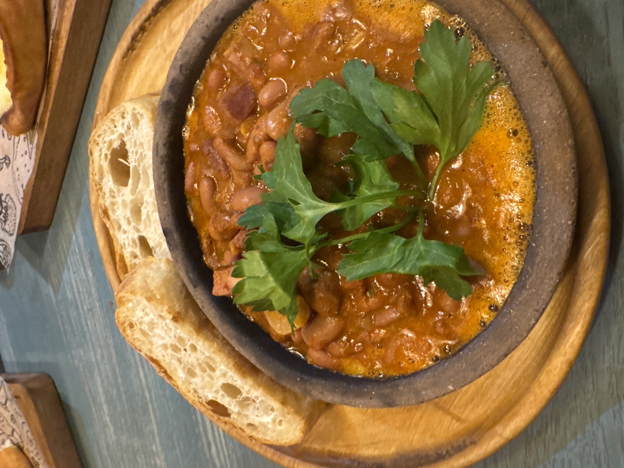 The image shows a bowl of beans in tomato sauce, garnished with fresh parsley, served with two slices of bread. The bowl is dark gray and sits on a round wooden platter. The bread has a porous texture and appears toasted. The food is positioned on a light blue wooden surface. The setting implies a restaurant or dining establishment. The image doesn't explicitly provide the location in Izhevsk, Russia, and there's no specific information about the presence or actions of people.