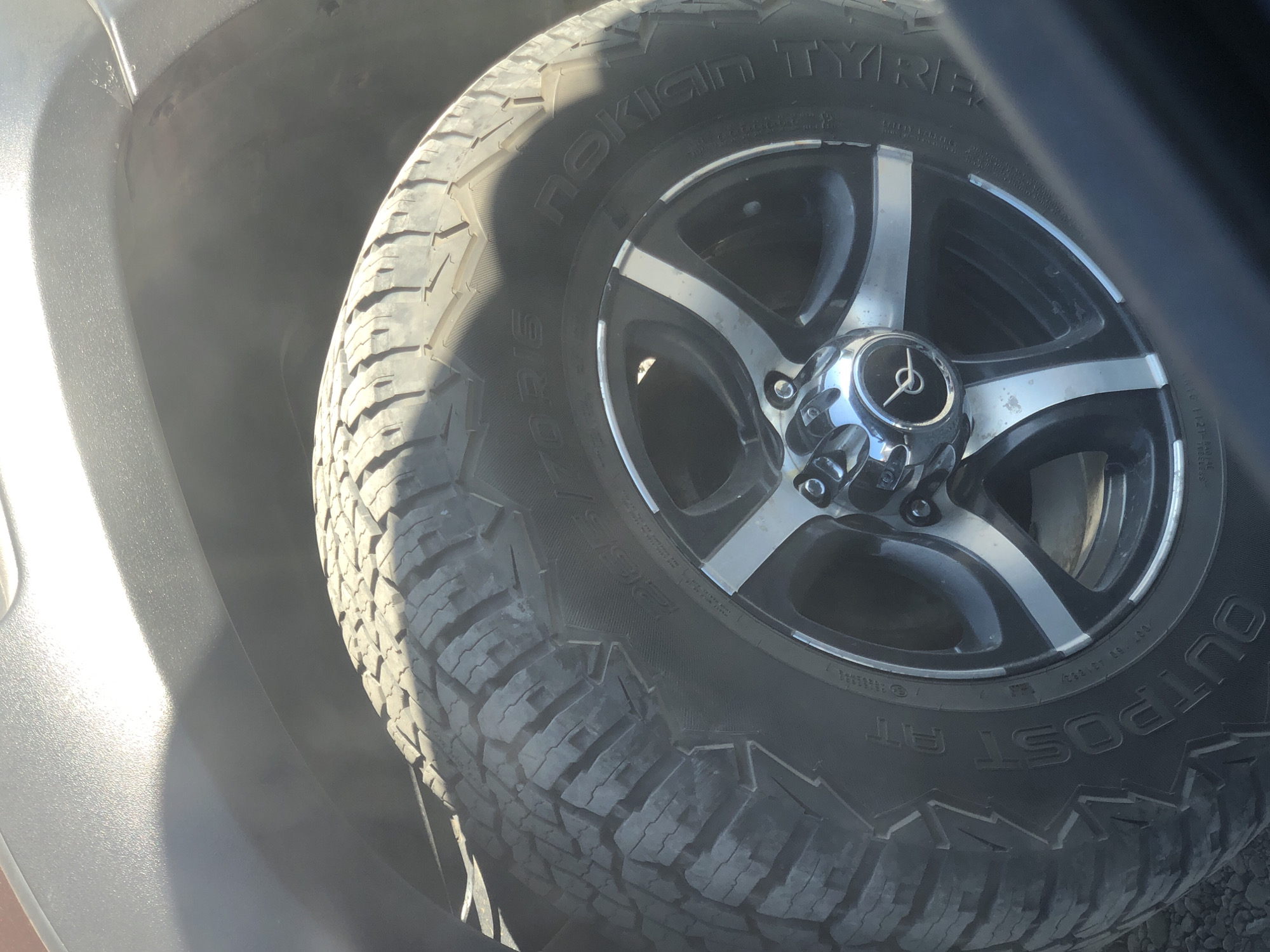 The image shows a close-up of a car's tire and wheel, likely taken in Petropavlovsk-Kamchatsky, Russia, as indicated by the prompt. The tire is an all-terrain type, with visible tread patterns and markings including "265/70R16" and "NOKIAN TYRES". The wheel appears to be an alloy rim with silver accents. The car's fender, which is dark gray and textured, is partially visible in the upper left portion of the frame. Sunlight is casting a shadow and glare across the top of the fender. The ground beneath the tire is covered in small stones or gravel. No people are visible in the image.