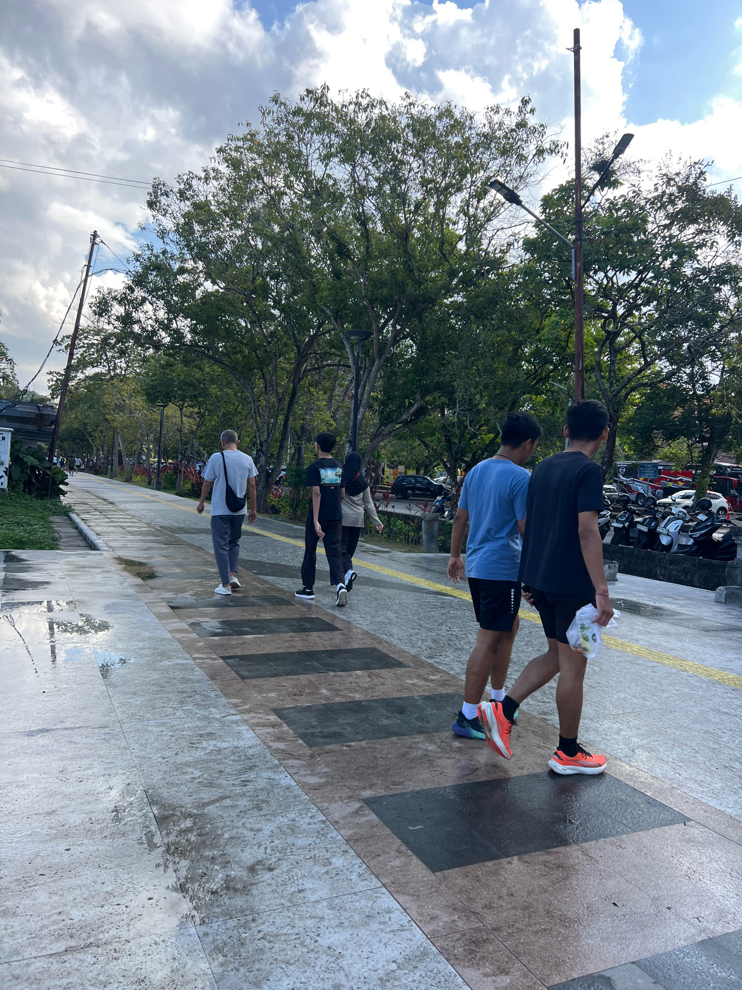 In Denpasar, Indonesia, a paved pathway in a park setting features several individuals walking. In the foreground, two young men walk together. The man on the left wears a blue t-shirt and black shorts, while the man on the right wears a black t-shirt and black shorts, carrying a clear plastic bag with some items inside. Behind them, a man with graying hair walks away from the camera, wearing a gray t-shirt and dark pants, with a black bag slung across his shoulder. Further up the path, another individual walks in the distance, wearing a black shirt and dark pants. The pathway is lined with trees and features streetlights. The ground appears wet, with reflections visible on the pavement, suggesting recent rain. In the background, a row of motorcycles and some parked vehicles are visible, along with more trees and overhead wires. The sky is partly cloudy with patches of blue.
