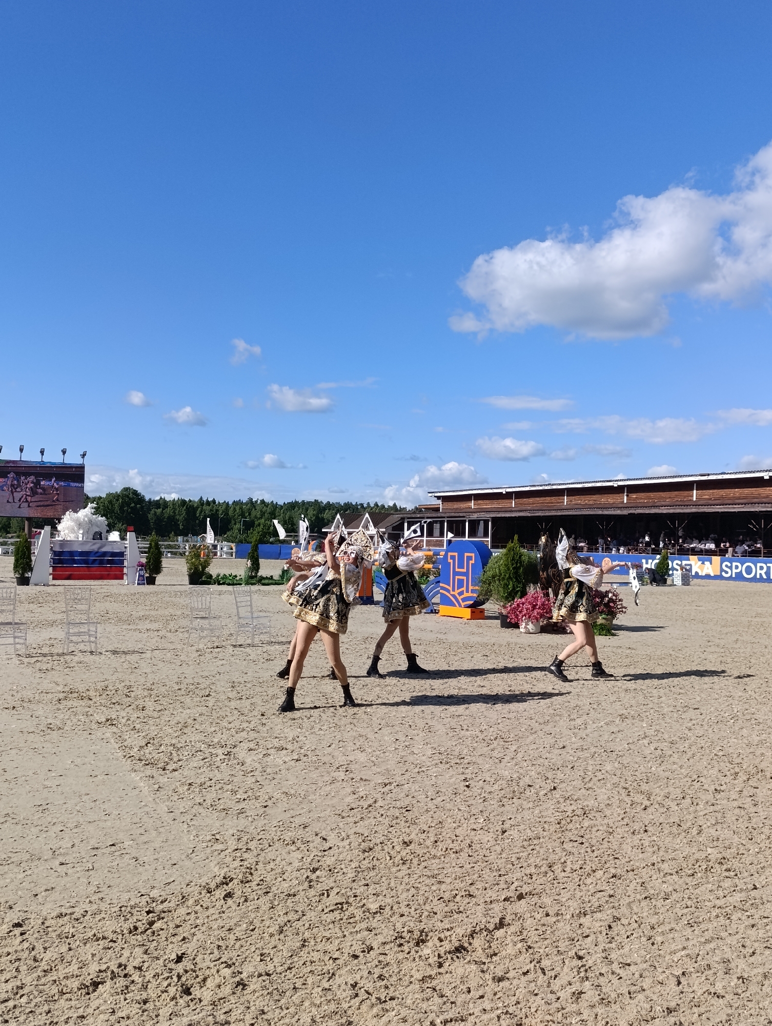 Three performers in elaborate gold and black costumes with headdresses are dancing on a sand arena, likely at a horse show or equestrian event in Ivanovskoe, Russia. The performers are positioned from left to right. The leftmost performer is turned away from the camera, while the middle and rightmost performers face more towards the viewer. They are engaged in a synchronized dance routine, with outstretched arms. The arena is sandy with tire tracks visible. In the background, there is a large building with a wooden facade, tents, trees, and a clear blue sky with scattered white clouds. A large screen displays an image of a horse competition. Several white chairs are set up in the foreground. The overall scene suggests a professional event with entertainment.