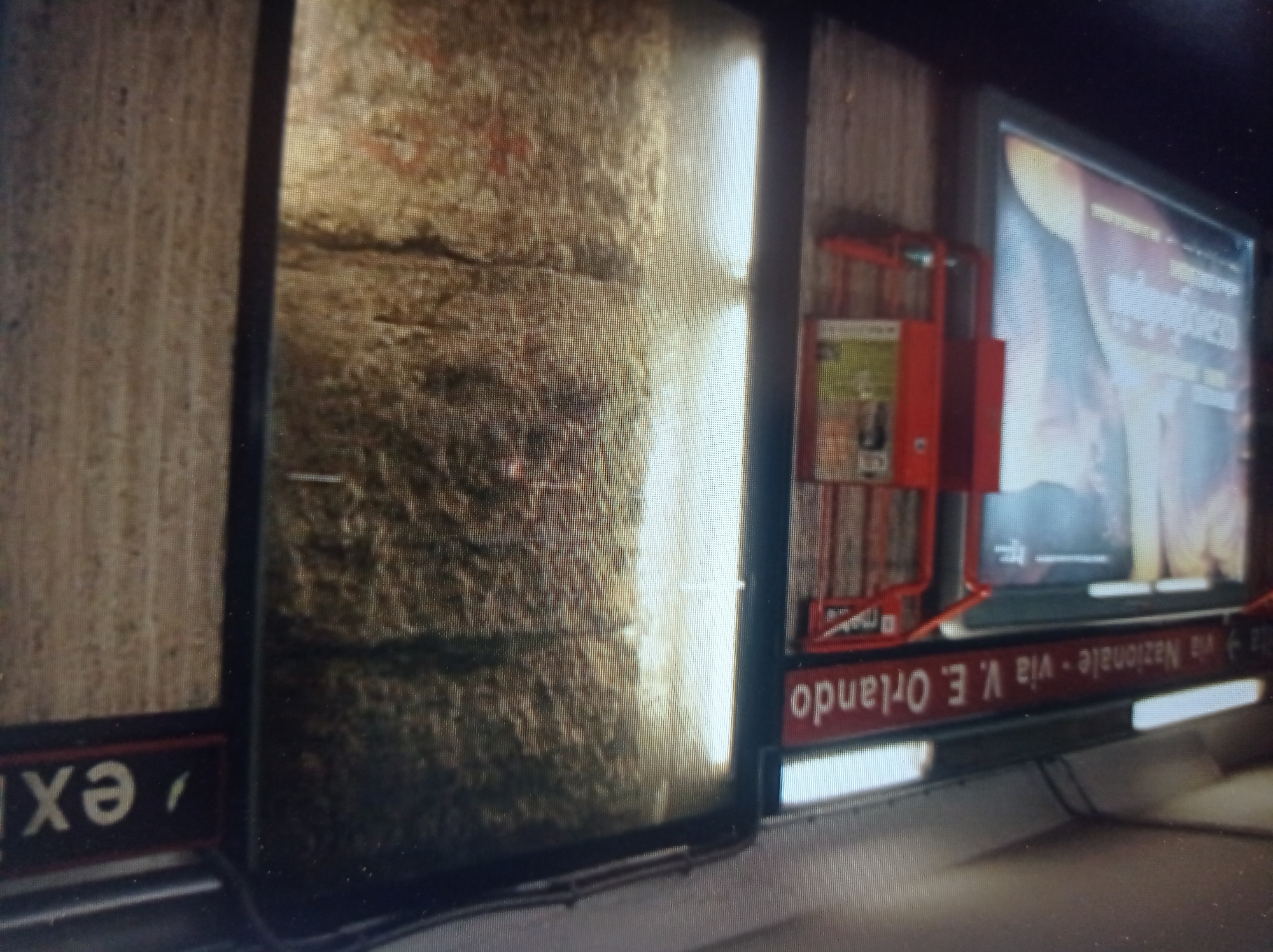 The image depicts a subway station or a similar underground passage. A red sign on the left reads "via Nazionale - via V. E. Orlando". To the right of this sign, a large illuminated advertisement is displayed. Further to the right, there is a large, textured wall and a red sign that says "exit". The overall scene appears to be dimly lit, with the advertisements and exit sign providing some illumination. There are no people or active interactions visible in the image. The environment suggests an urban public transit setting. The provided information about the city of Mukachevo, Ukraine is not visually supported by the image content.