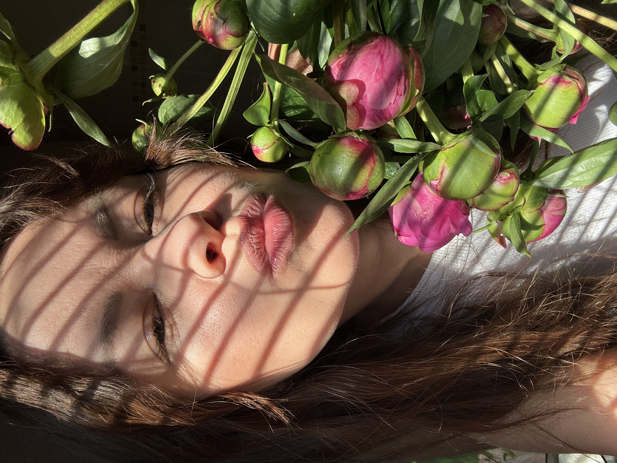 A young woman with brown hair is shown in a close-up portrait. She is leaning forward, puckering her lips, and holding a bouquet of pink peonies. The sunlight casts striped shadows across her face. The image is taken in Chaikovsky, Russia.