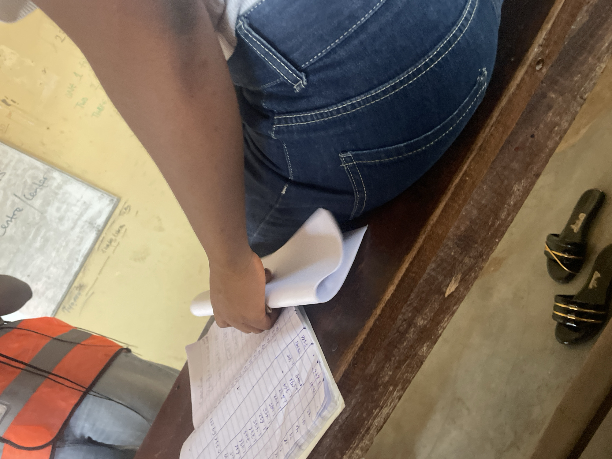The image depicts a close-up view of a person's legs and arm, seated at a wooden desk in what appears to be a classroom or administrative setting in Lokoja, Nigeria. The person is wearing blue denim jeans and a grey shirt, partially visible. Their right arm is bent, holding a stack of white paper. In front of them, an open notebook with handwritten entries, including numbers and what look like names or labels, is laid out on the desk. The notebook is ruled with columns. The background features a yellow wall with some writing on it, including the words "Centre" and "center", and possibly some numbers and symbols. A whiteboard or noticeboard is also visible in the background, with more handwritten text. The desk itself is made of dark, worn wood. In the lower part of the image, a pair of black sandals with gold accents are placed on the floor. The overall scene suggests an indoor environment where administrative tasks or data entry are being performed.