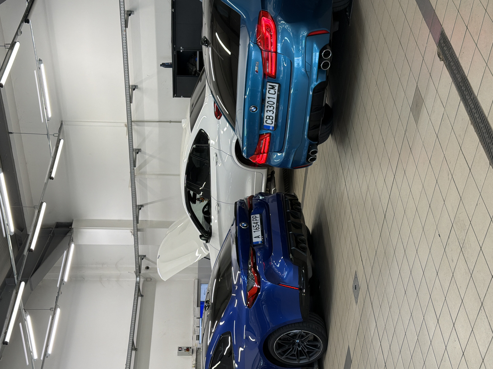 Three BMW cars are parked in what appears to be a service bay or showroom. The car in the foreground on the left is a blue BMW M4 coupe, with its rear end facing the camera. To its right and slightly behind is a teal blue BMW M5 sedan, also with its rear facing the camera. Further back and to the left is a white BMW SUV with its hood open. The environment is a well-lit indoor space with tiled flooring and overhead fluorescent lighting. A black storage cabinet is visible on the right side of the frame. The license plate on the blue BMW M4 reads "BG A 1654 PP". The license plate on the teal BMW M5 reads "BG CB 3301 CM". The scene is static with no people visible. Based on the license plates with the "BG" prefix, the location is in Bulgaria. The presence of specific car models and the nature of the environment suggest a car dealership or a specialized automotive service center.
