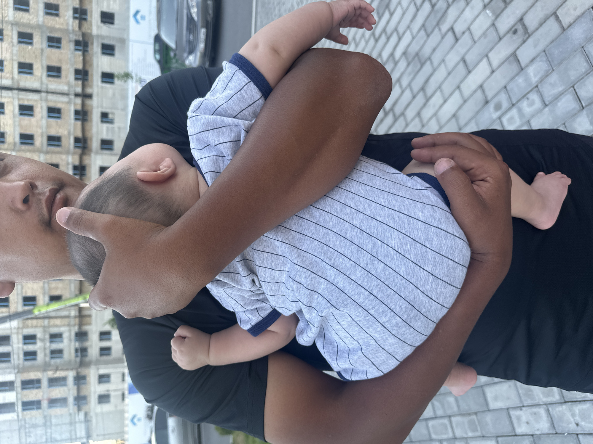 A man holds a baby wrapped in his arms. The baby is wearing a gray onesie with thin black stripes and a blue trim. The man is wearing a black t-shirt. The background shows a building under construction and a street with cars. The ground is paved with gray interlocking bricks. The scene appears to be an outdoor location in the city of Manas, Kyrgyzstan.