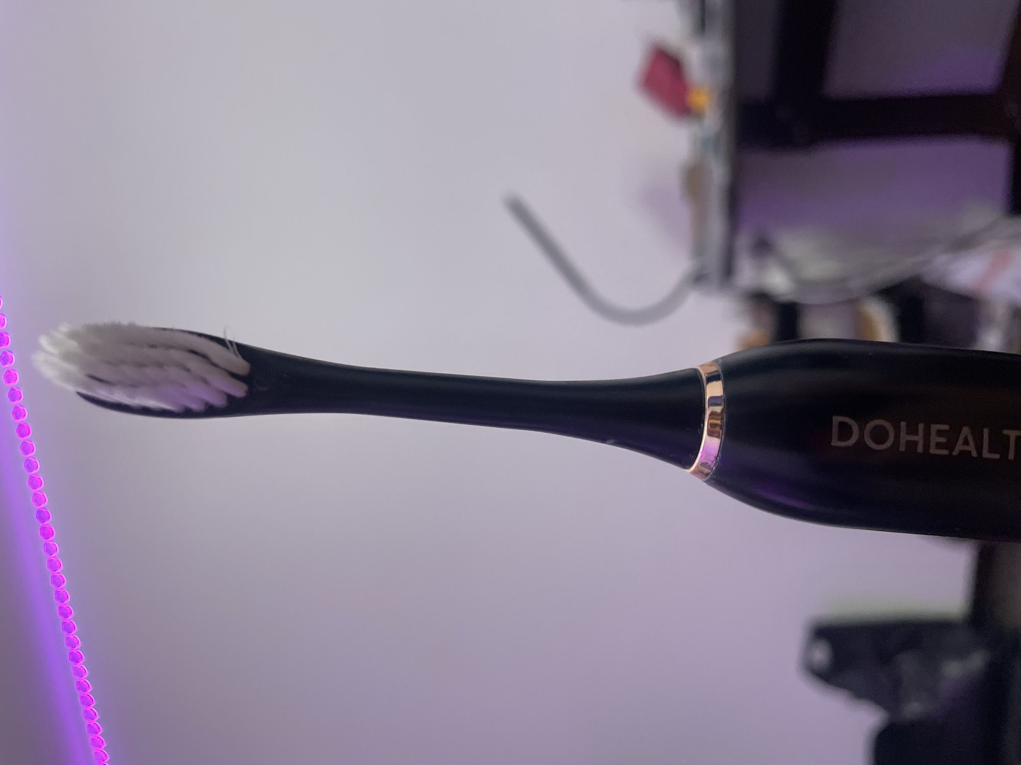 A black electric toothbrush with white bristles and a gold ring near the brush head is held in the foreground. The brand name "DOHEALTH" is printed vertically in white text on the toothbrush handle. In the background, a purple LED light strip runs across the top of the image, and a blurred desk with various items, including a pink object and a lamp, is visible. The scene is indoors, with a plain wall as the backdrop. The exact location in Kokkorevo, Russia cannot be determined from the image.