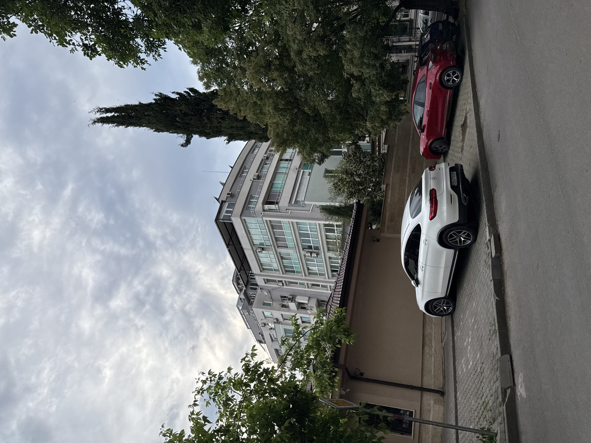 The image depicts a street scene in Stara Zagora, Bulgaria. The view is angled upwards, capturing a multi-story building with numerous windows and air conditioning units. The sky is overcast with cloudy conditions. Trees frame the upper portion of the image, with one particularly tall, slender conifer.
On the right side of the frame, two parked cars are visible along the curb: a white SUV, likely a Mercedes-Benz GLC model (registration number is blurred, but the characters CB 6710 XP are somewhat visible) and a red hatchback. The street surface appears to be paved, and the sidewalk is constructed of interlocking pavers. The surrounding environment includes other buildings and foliage, typical of an urban setting. No people are visible in the image.