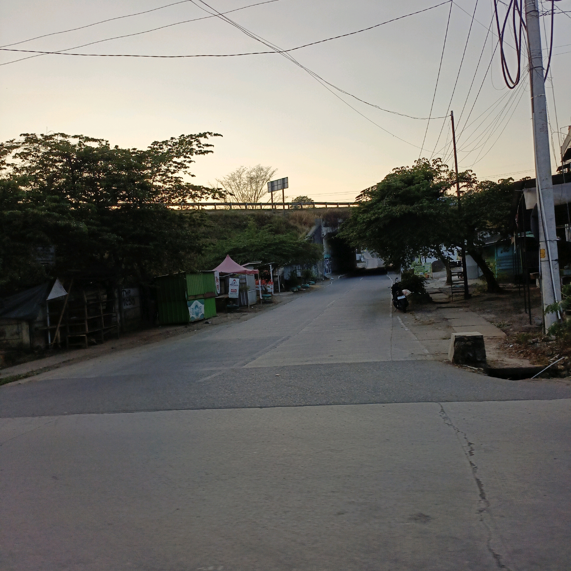The image depicts a street scene in Salatiga, Indonesia, likely during the late afternoon or early evening, indicated by the soft, warm light and the pale sky. The perspective is from the ground, looking down a paved road that curves to the right and disappears under a bridge or overpass.
On the left side of the road, there are several small, makeshift structures, possibly stalls or shops. One has a red tent, and another is a green shipping container. Behind these structures, dense green foliage and trees line the embankment of the overpass. A few signs are visible, but their text is mostly illegible.
The road itself appears to be in fair condition, with some visible cracks and a distinct line marking the center or lane division. On the right side of the road, more trees and vegetation are present. A concrete structure, possibly a drainage cover or a bench, is visible near the edge of the road. A parked motorcycle is also present on the right side of the road, near the curb.
Overhead, a network of electrical wires crisscrosses the sky, supported by poles, typical of urban or semi-urban environments in Indonesia. The overpass structure is made of concrete and appears to be elevated, with a road or railway passing over it. The overall impression is a somewhat cluttered, functional street with elements of everyday life and infrastructure. The specific location within Salatiga is not identifiable from the image alone.