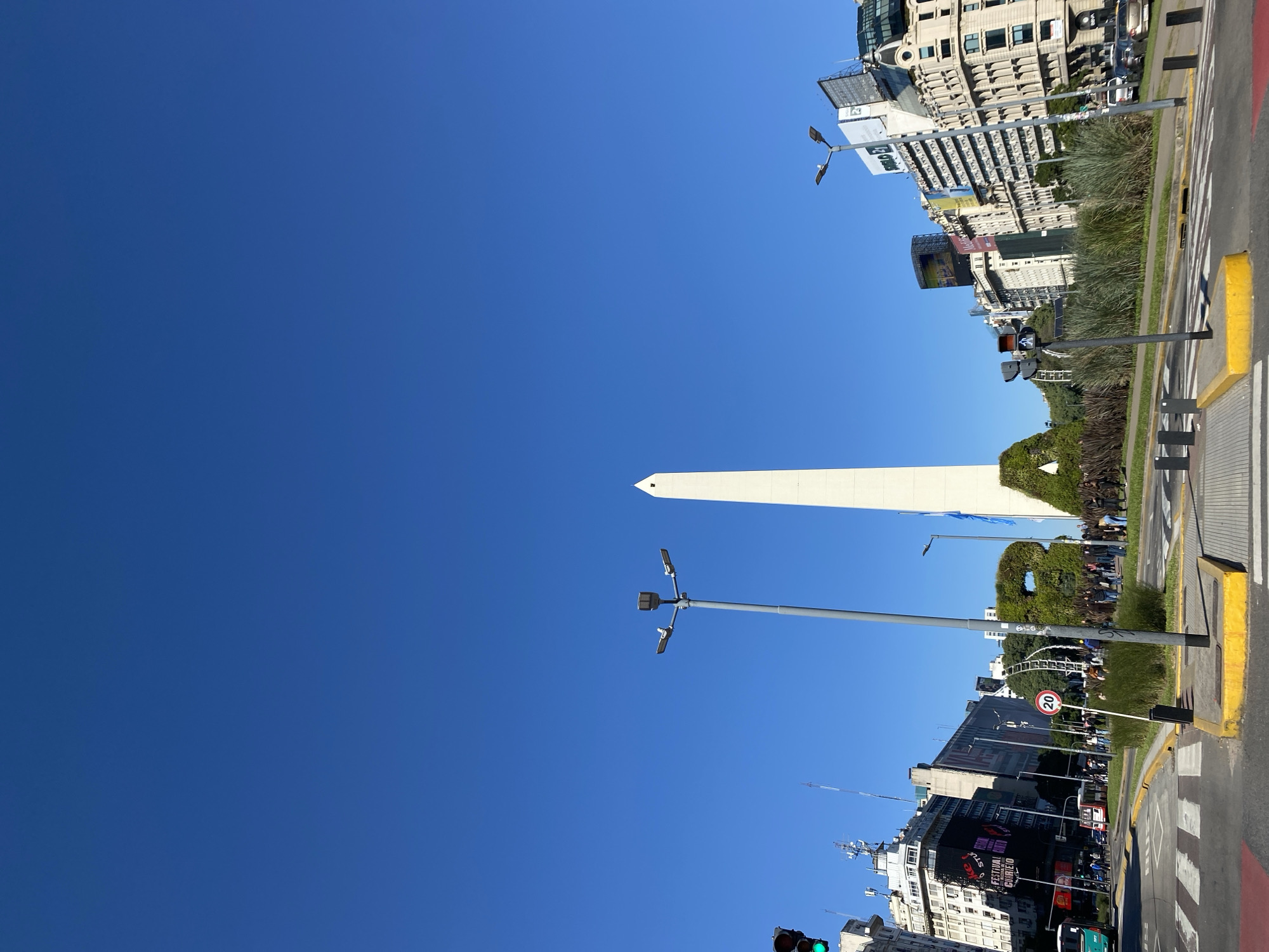 The image depicts the Obelisco de Buenos Aires, a historical monument located in the city of Buenos Aires, Argentina. The obelisk is a tall, white structure, pointed at the top, situated at the center of Plaza de la República. The view is from below, looking up toward the monument. The sky is clear and blue. Street lamps are visible. Buildings of different heights and architectural styles, some with advertisements and signs, line the streets. Pedestrian crossing stripes mark roadways with yellow curbs and planted greenery. The image captures a sunny day in the city.