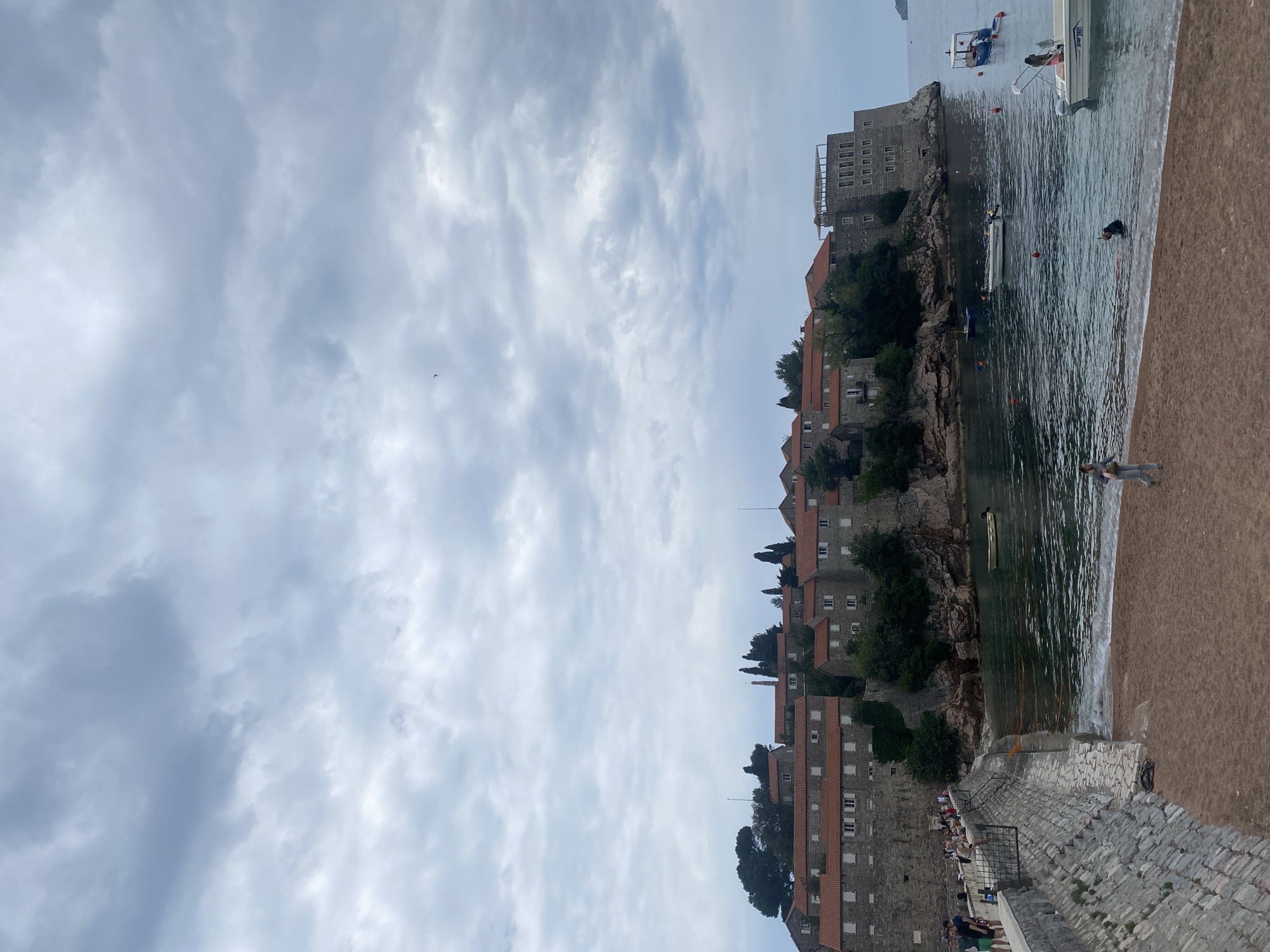 The image captures a serene view of Sveti Stefan, Montenegro. The scene features a coastline with a series of buildings constructed with grey stone walls and orange tiled roofs, perched along a cliff. Buildings rise incrementally up the cliff-side, interspersed with verdant trees. The water meets the shore, with small boats floating offshore and people wading in the water. In the foreground, a sandy beach meets a stone pathway where people are sitting and walking. The sky is overcast with a dense layer of clouds.