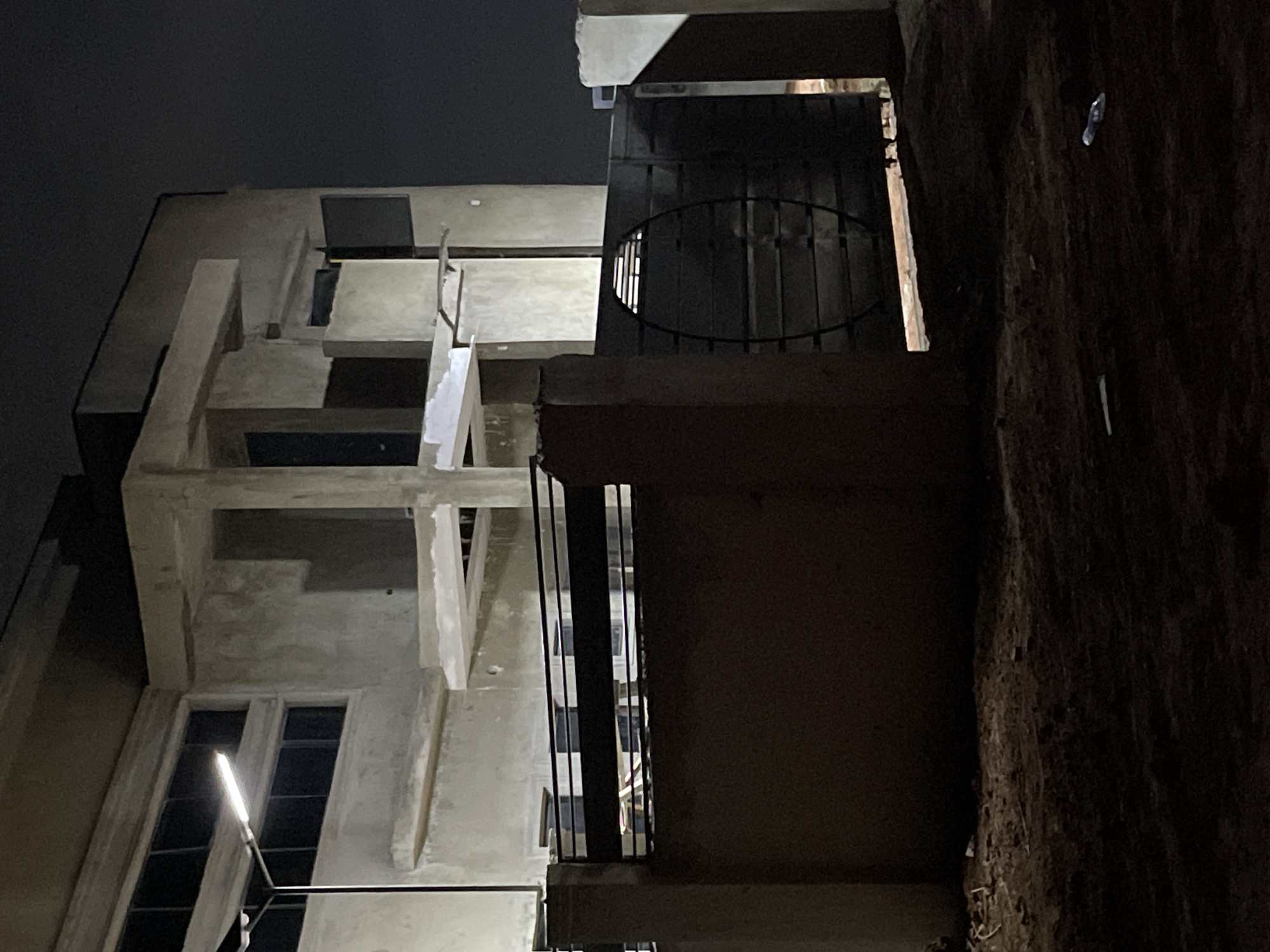 The image depicts a low-angle night view of buildings under construction in Angwan Sayawa, Nigeria. The structure on the left is a light-colored, multi-story building with exposed concrete framework and unfinished windows. One window on the lower floor emits a bright light. To the right, a darker, seemingly older building or wall is visible, with a round metal railing or decorative element protruding from one of its levels. The sky is dark and overcast. The overall scene appears to be a contrast between the unfinished construction and a more established structure.