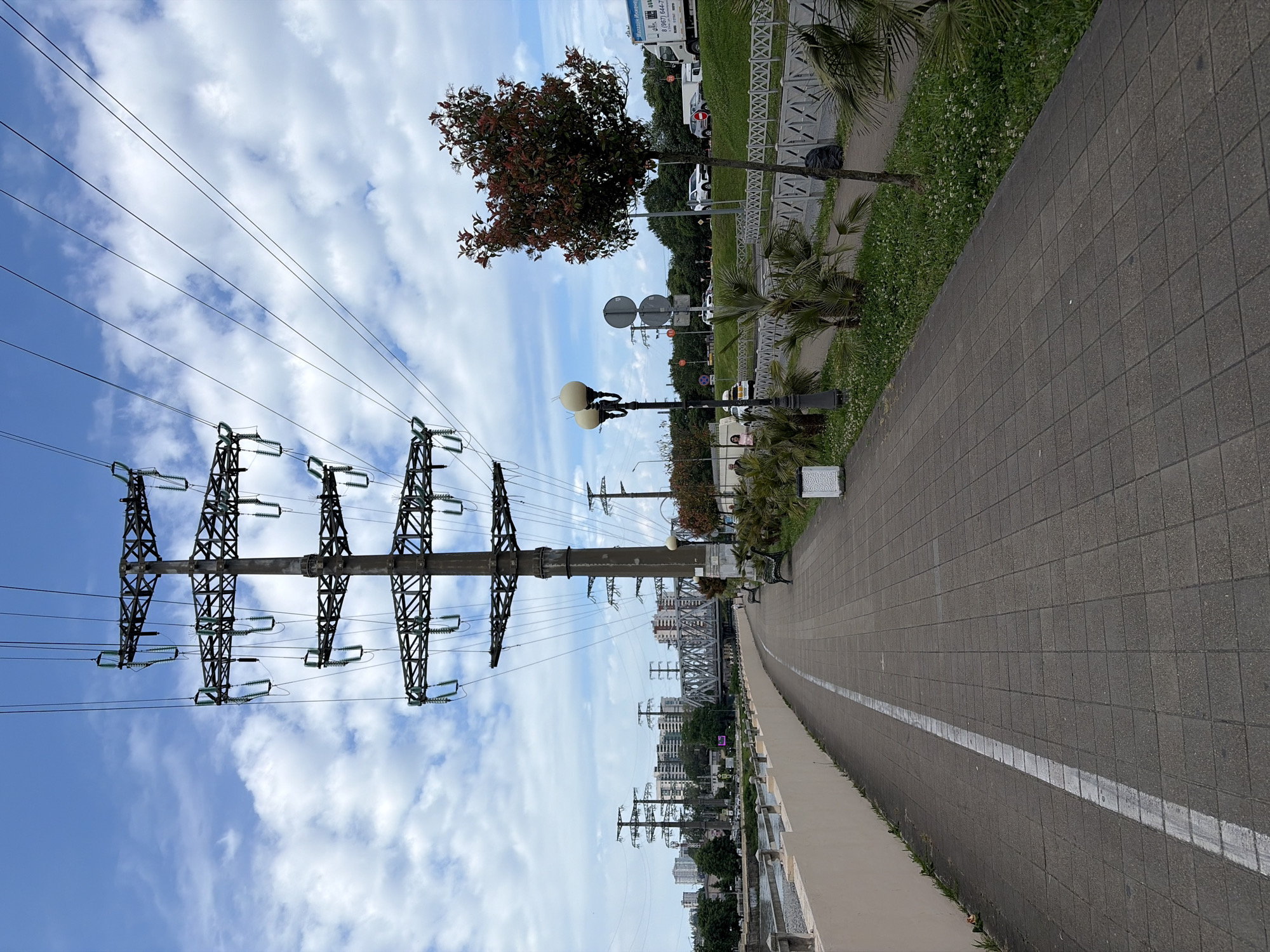 The image depicts a paved pedestrian walkway along a body of water, likely a river or sea channel, in Sochi, Russia. Tall electricity pylons with multiple crossbars and insulators dominate the scene, with power lines stretching across the frame against a partly cloudy sky. The walkway is bordered by a concrete barrier on the left and a grassy area with small trees and palm-like plants on the right.
In the background, a bridge, possibly a railway bridge, is visible, along with apartment buildings and other infrastructure. Several cars and a truck are present on a road beyond the grassy area. A street lamp with two globes stands near the walkway, and a park bench is visible further down. Two people, one standing and one walking, are present on the walkway. A trash can is located to the right of the walkway. The overall impression is an urban waterfront area with significant industrial infrastructure.