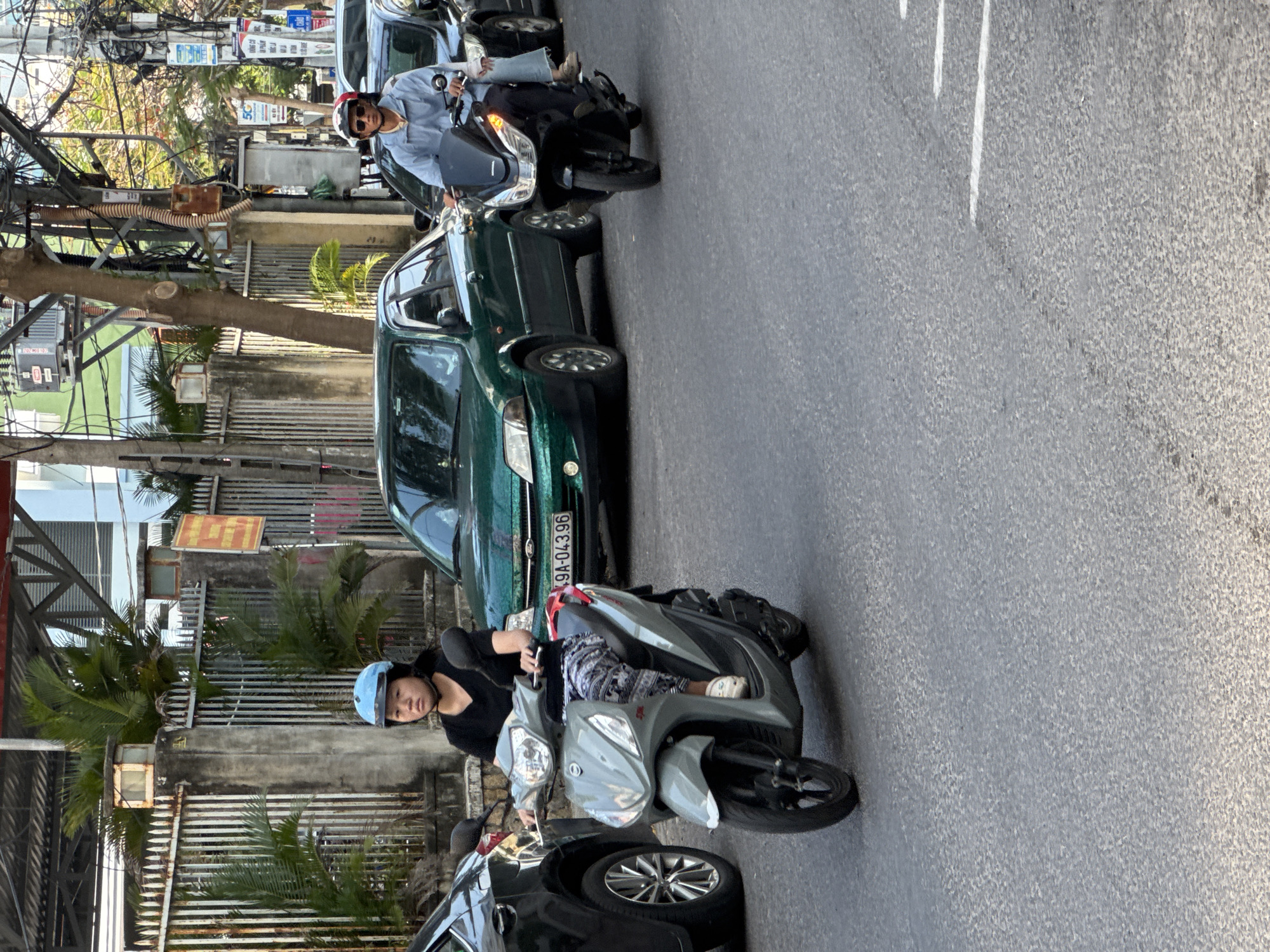 A young Vietnamese woman wearing a light blue helmet and a black shirt is riding a light gray scooter on a paved road. She is looking forward and appears to be in traffic. Her right leg is bent at the knee, with her white shoe visible. She is wearing patterned pants. In the background, a green car is visible, followed by a black car. Further in the background, there is a metal fence and tropical foliage. On the right side of the image, another scooter is being ridden by a man wearing a light blue shirt and sunglasses, with a passenger behind him. This scene is typical of urban traffic in Nha Trang, Vietnam, characterized by dense scooter and car traffic on asphalt roads, often with overhead power lines and adjacent sidewalks or buildings.