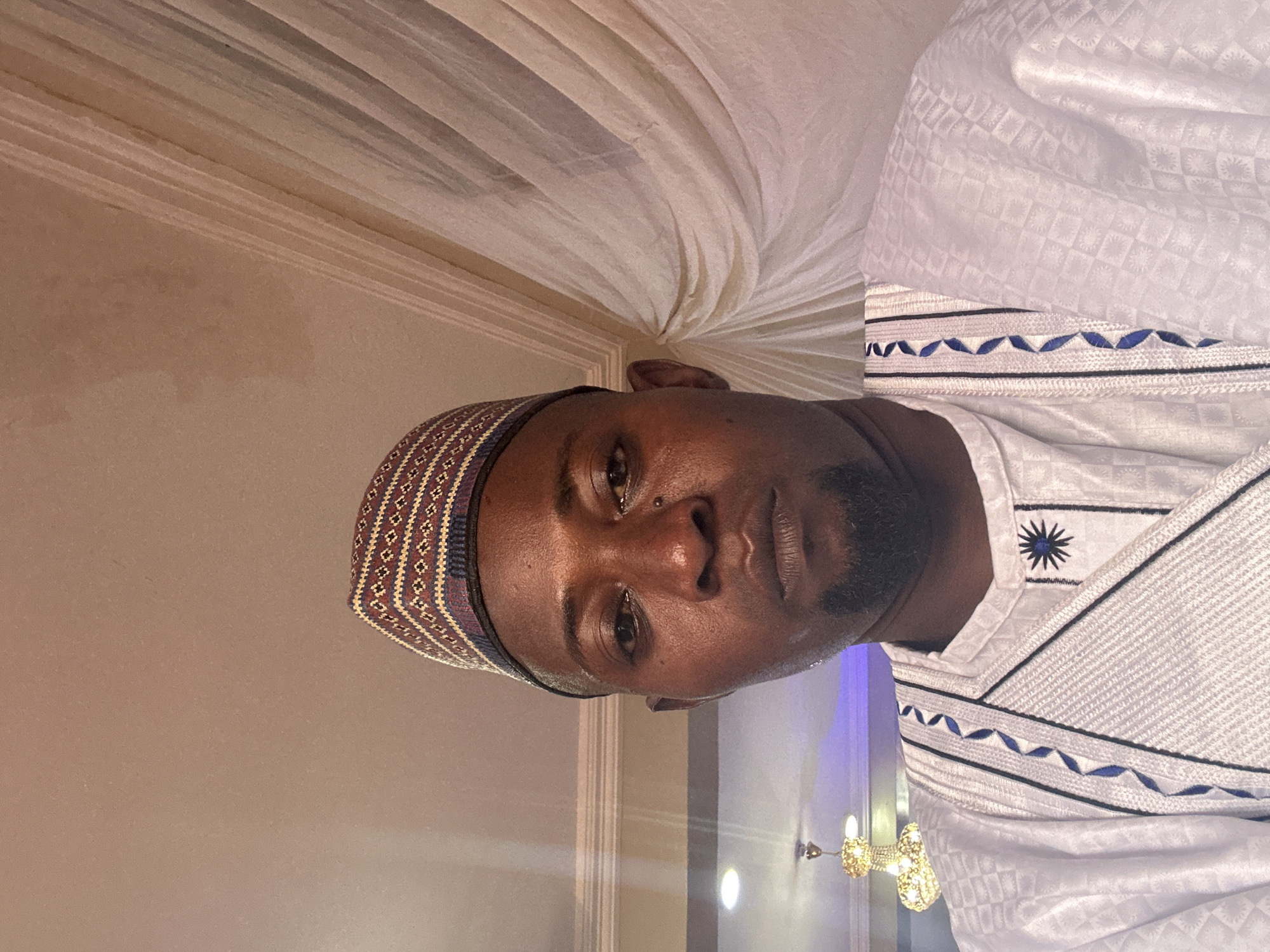 An image shows a man, likely of African descent, looking directly at the camera with a neutral expression. The image is oriented with the man's head tilted to the left. He has dark skin, a short beard, and a visible nose piercing. He wears a traditional kufi-style cap adorned with intricate geometric patterns in shades of red, gold, blue, and white. His attire is a white garment, possibly a dashiki or thobe, featuring a subtle embossed or woven pattern of squares and diamonds. The garment has distinctive blue and black embroidery along the neckline and chest, including wavy lines and a star-like motif.
The setting appears to be indoors, possibly a formal event space, hall, or a decorated room. The background includes light-colored, draped fabric on the ceiling, and hints of warm-toned walls. In the lower right corner, a blurry, ornate light fixture resembling a small chandelier is visible, suggesting artificial lighting. The overall lighting is warm, contributing to an indoor, possibly evening, ambiance. There is no visible text in the image.