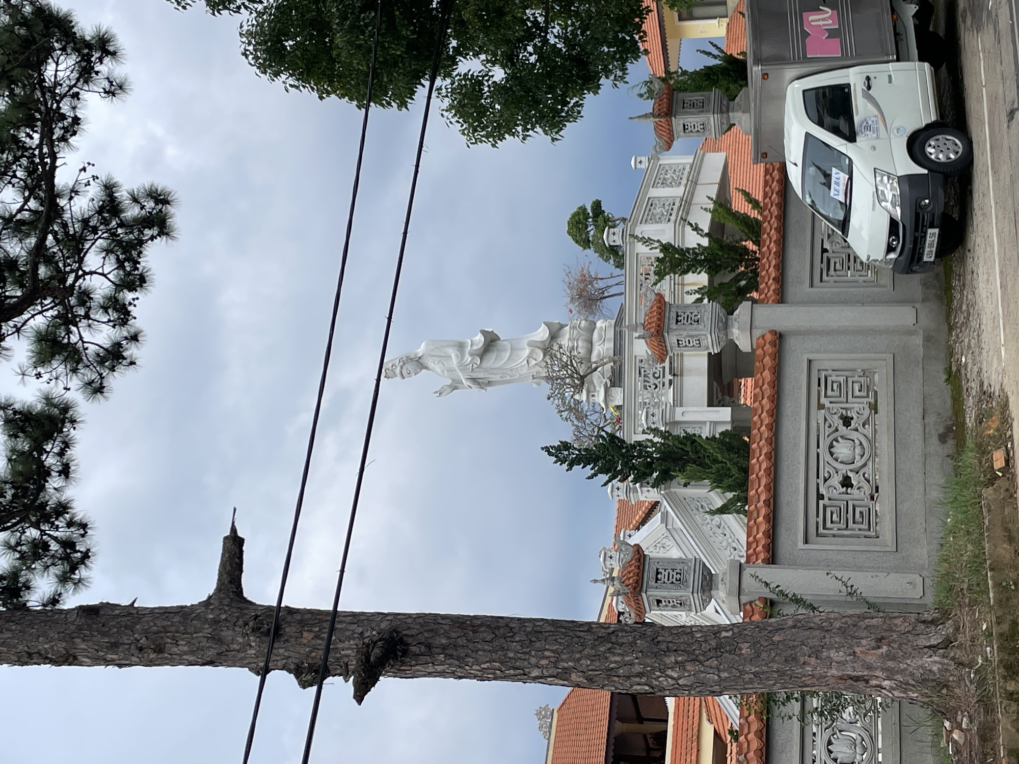 In Lâm Viên - Đà Lạt, Vietnam, a white delivery truck with a silver cargo box is parked on the side of a road. The truck has a license plate reading "49A-984.56" and a pink logo on the cargo box. It is parked in front of a gray stone wall with ornate carvings and an orange tiled roof. Behind the wall, a large white statue of a deity stands on a multi-tiered platform with decorative lanterns and greenery. A tall pine tree with thick branches is in the foreground to the left of the frame, and a lush green tree is on the right. Overhead, power lines cross the overcast sky, which is filled with dark, dramatic clouds. The scene appears to be a public area, possibly a temple or cultural site.