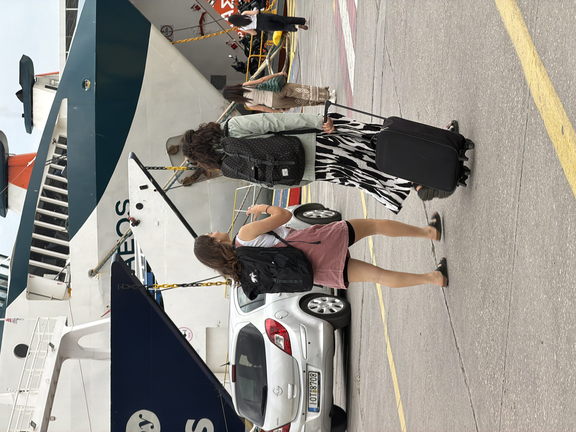 A medium shot of a dock in Piraeus, Greece, focuses on pedestrians and a small car in front of a docked ship. A woman in a red dress is holding her phone up and to the side, as if taking a picture of the ship. She has on a white undershirt and a black backpack. Her position puts her in front of a white Opel Corsa hatchback with the license plate IOT 8708. To the left of the car is the ship. The visible letters "EOS" are emblazoned on its side. Two other women are walking away from the ship, towards the right of the frame. The first woman is wearing a light green jacket, a backpack with black and white polka dots, and a black and white zebra-striped skirt. She is pulling a black rolling suitcase. The second woman is wearing a tan shirt and a tan skirt with brown patterns. She is also pulling a rolling suitcase. The foreground is cracked concrete. A yellow painted line is visible along the right edge of the frame.