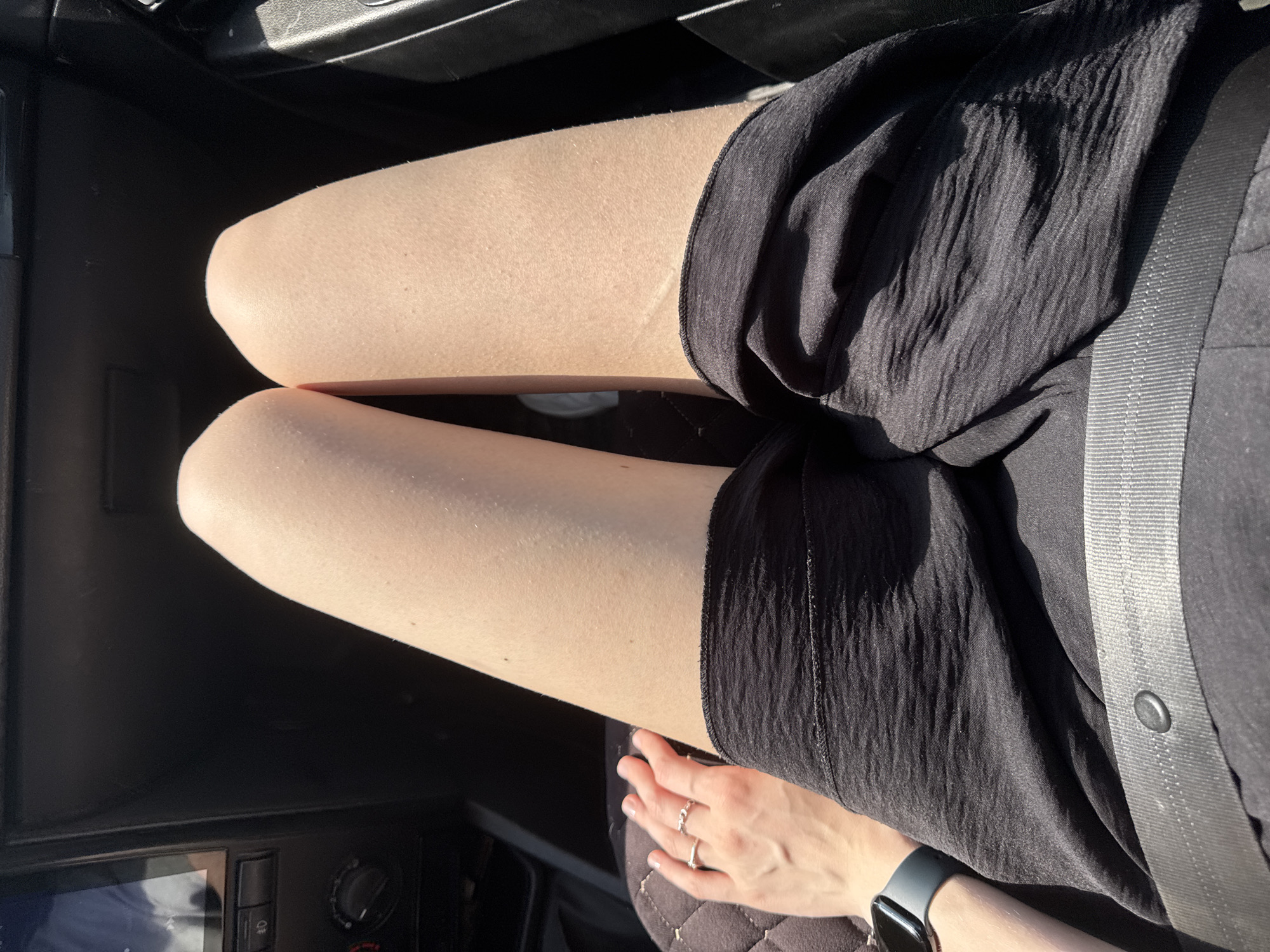 The image shows a person seated in a car, wearing black shorts, with a seatbelt fastened across their body. The person's hands are visible on their lap, adorned with rings and an Apple Watch. The interior of the car is partially visible, including the dashboard and center console. The location is unknown.