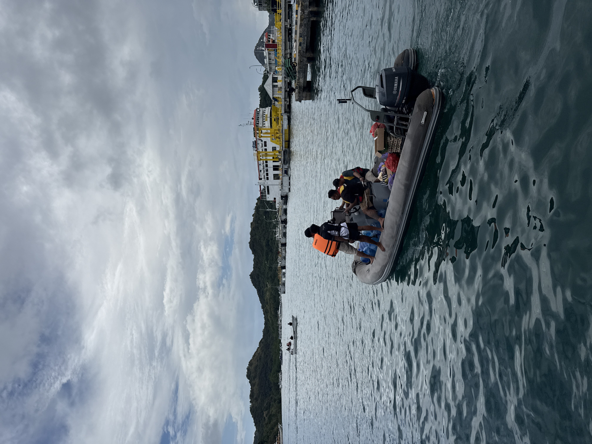 The image shows a grey inflatable boat with a Yamaha outboard motor in the water near Labuan Bajo, Indonesia. The boat is carrying multiple people and supplies. Some passengers are wearing orange life vests. Behind the boat, there is a yellow and white ferry docked at a pier. Green hills and buildings are visible in the background. The sky is cloudy. A smaller boat is in the water further away.