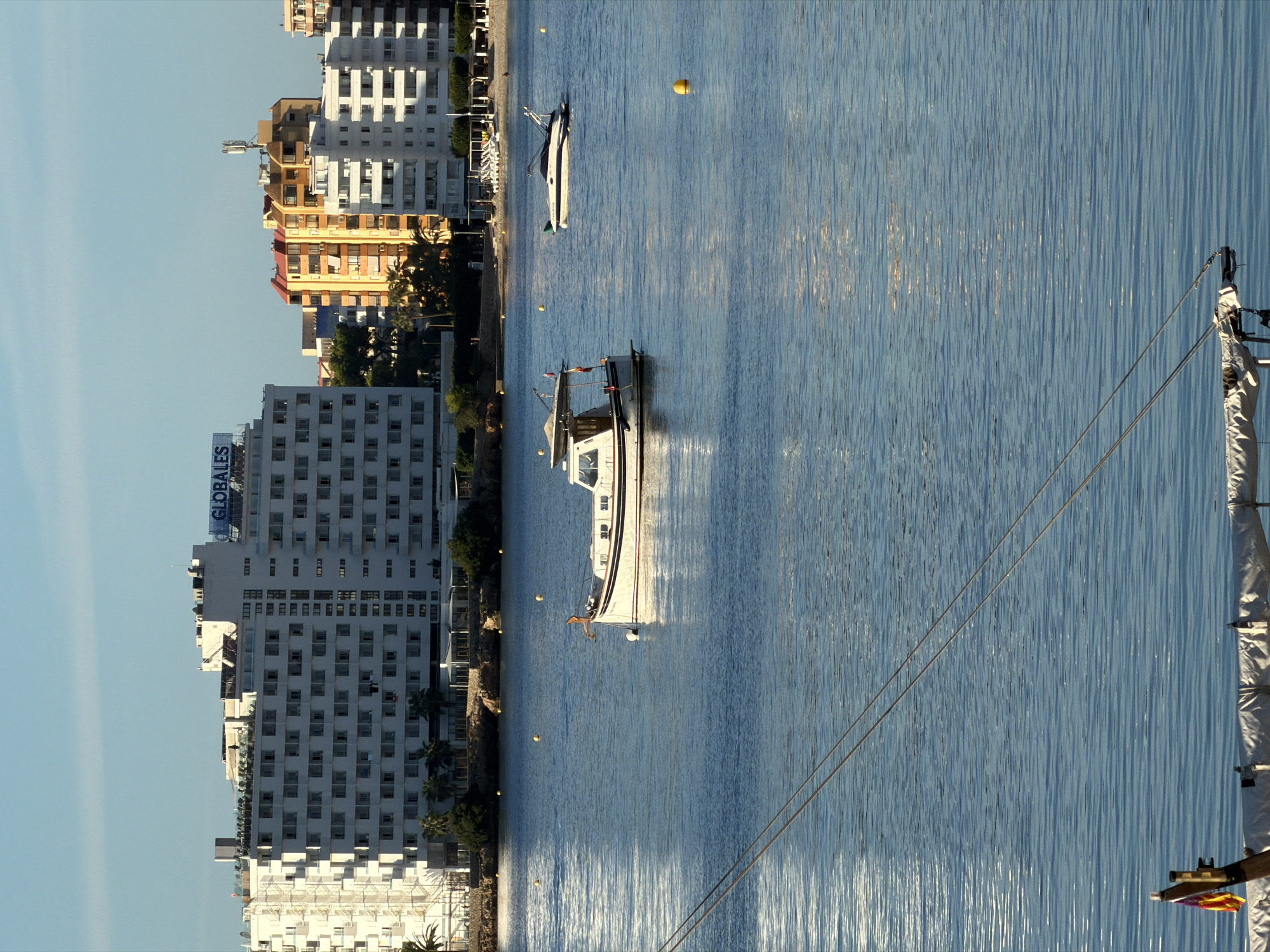 The image depicts a scenic view of the waterfront in Palmanova, Spain, with a clear blue sky and calm sea. In the foreground, part of a sailboat is visible with a folded sail and rigging. Two other boats are anchored in the water: a white motor yacht with dark trim and an orange anchor at its bow, and a smaller white boat with a canopy. In the middle ground, a sandy beach with beach chairs and umbrellas can be seen, backed by a line of trees and then a series of multi-story buildings. The largest building, on the left, is a white hotel with the blue "GLOBALES" logo on its roof. To its right, a cluster of brightly colored buildings, including orange and yellow structures, rise against the sky. Yellow buoys are scattered in the water. The overall scene conveys a peaceful, sunny atmosphere typical of a coastal resort town.