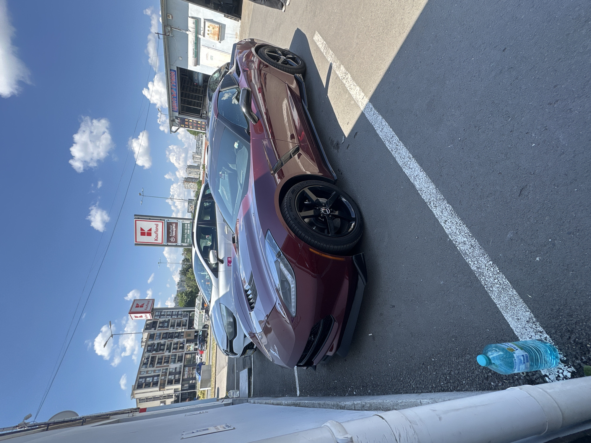 The image shows a parked burgundy Chevrolet Corvette in a parking lot on a sunny day. The car is the primary subject, with its sleek lines and sporty design prominently displayed. The setting is an outdoor parking area, likely in an urban or commercial zone, with buildings and signs visible in the background.
In the background, there are several signs for "Kaufland," a supermarket chain, suggesting the location is near a shopping center. A white car is parked next to the Corvette. The sky is blue with fluffy white clouds, indicating fair weather. The sun is casting shadows, suggesting it is daytime. There is also a large plastic bottle of water lying on the ground near a white line painted on the asphalt.
The image is captured from a low angle, emphasizing the car's presence. There are no people clearly visible in the frame. The overall mood is casual, reflecting a typical day in a parking lot. Text visible on the signs includes "Kaufland" and some Bulgarian text, possibly related to store hours or departments. The tire of the Corvette also has "KUMHO" printed on it, indicating the tire brand.