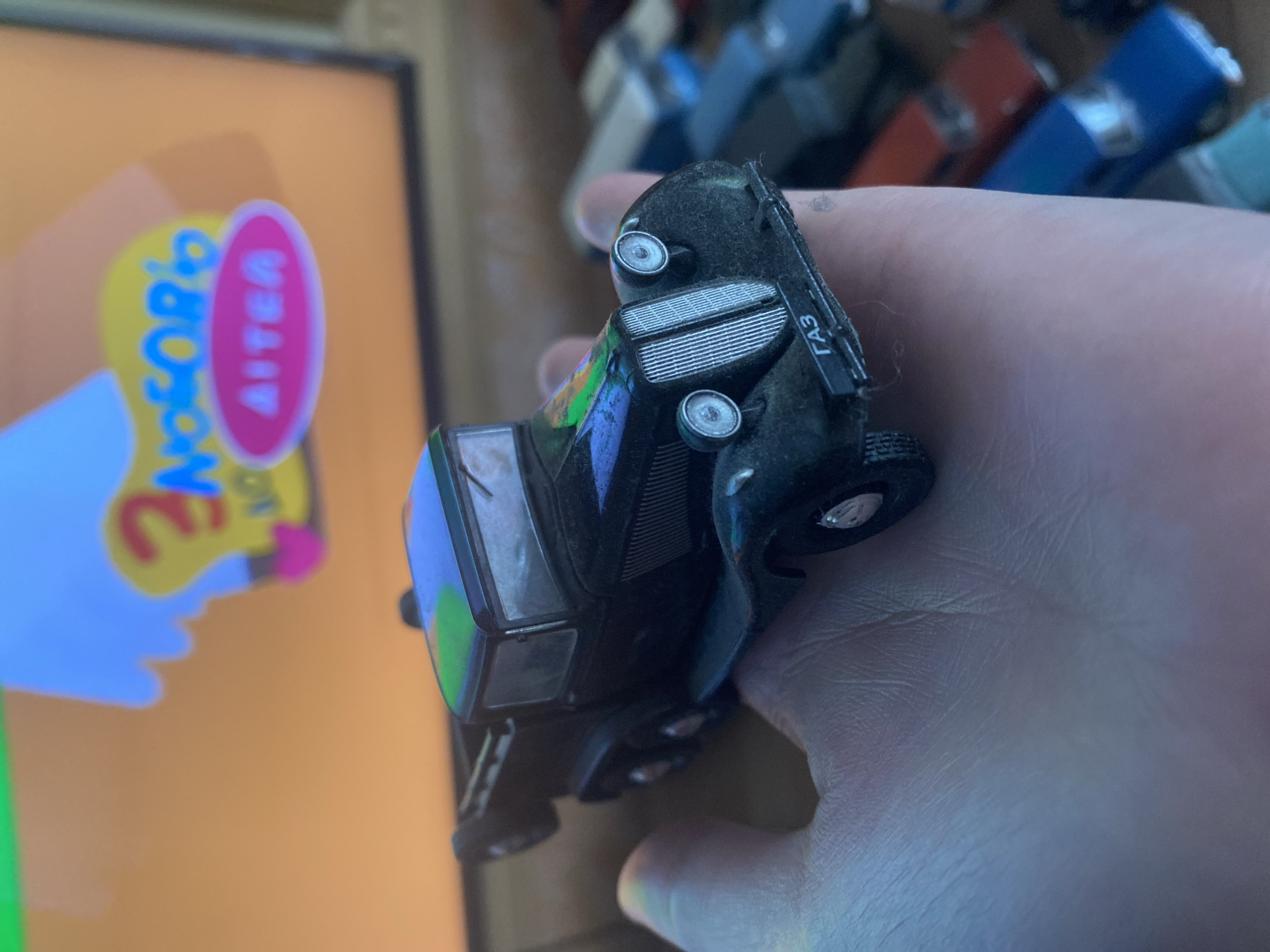 The image shows a hand holding a black toy truck with the letters "ГАЗ" on the front bumper. The truck has a detailed grill, headlights, and a visible cab with windshield wipers. In the background, a television screen displays a cartoon with a yellow character and blue text. Several other toy cars are visible in the lower portion of the image, including red and blue models. The setting appears to be indoors, likely a living room or play area. There are no people or recognizable landmarks present to indicate a specific location in a city.