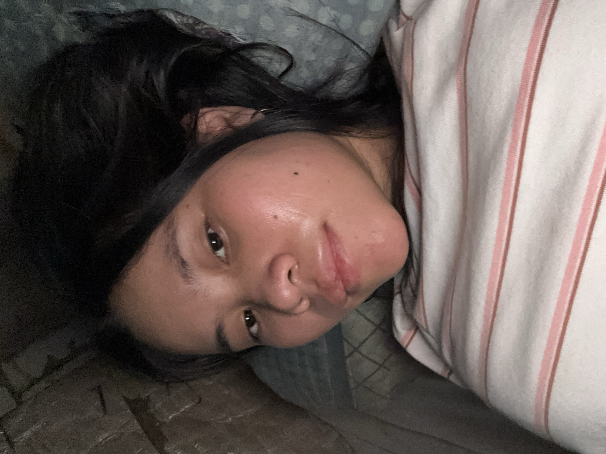 A young woman is lying down, her face is captured in a close-up shot. She has dark hair, brown eyes, and appears to be in a dimly lit room. She is wearing a striped shirt. The background shows a textured surface on the left and a patterned fabric on the right. The image is a selfie taken from a high angle, suggesting the woman is holding the camera. The overall scene is intimate and casual, possibly taken in a bedroom. East Jakarta, Indonesia, is not depicted or implied in the image.