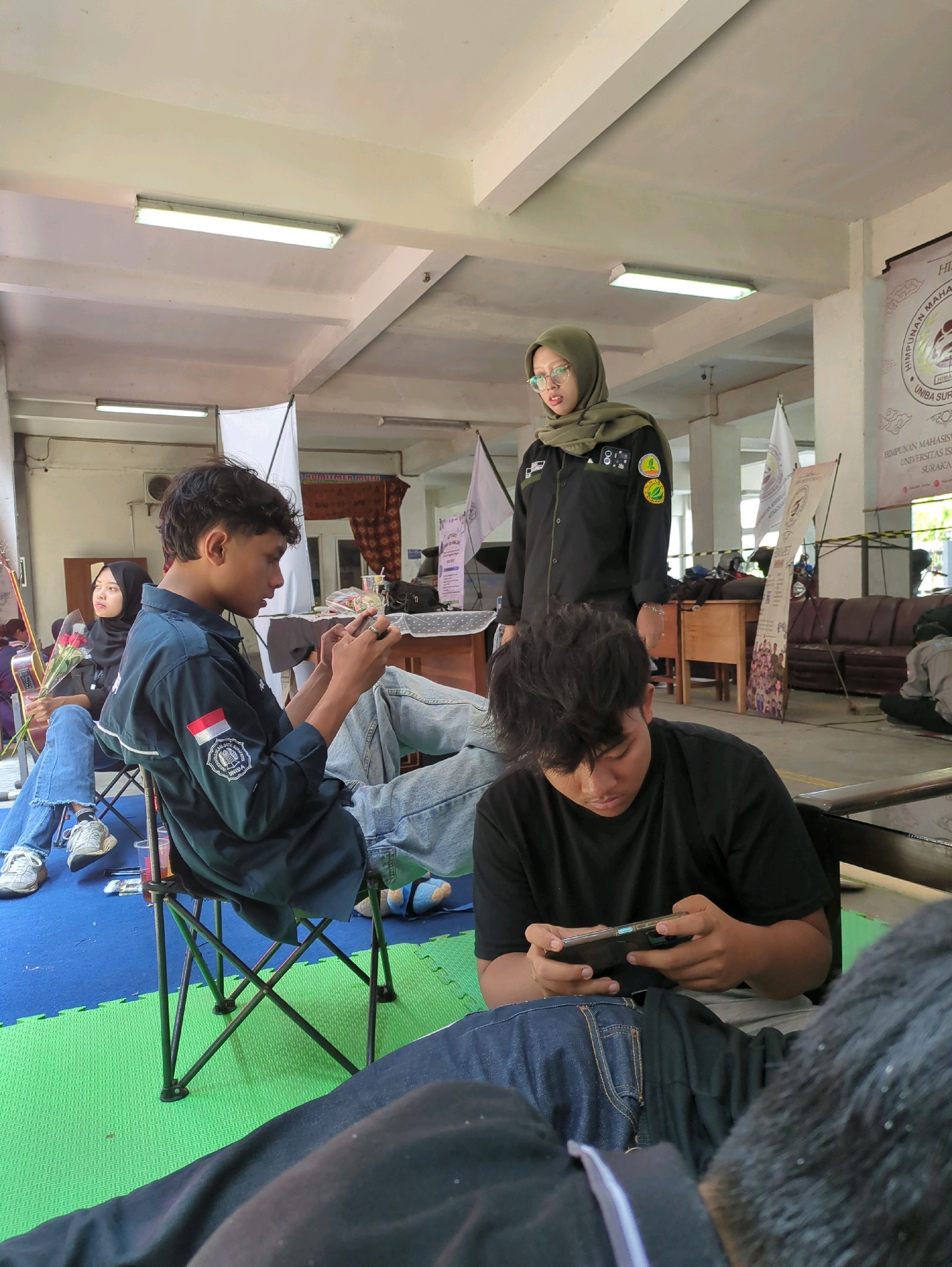 A group of young people are gathered in a room that appears to be an indoor event space or classroom at an educational institution in Surakarta, Indonesia. The room has a high ceiling with visible fluorescent lighting and exposed beams. The walls are a light color, and banners with text and logos are displayed, suggesting an organized event or exhibition.
In the foreground, two young men are engrossed in their mobile phones. One, wearing a black t-shirt, is seated and looking down at his device, held horizontally in his hands. The other, wearing a dark blue uniform shirt with patches on the sleeve, is also seated and actively playing on his phone. He is positioned in a foldable chair with a dark frame. Behind him, to the left, a young woman wearing a black hijab and blue jeans is also seated, holding a bouquet of red roses and what appears to be a part of a musical instrument.
A young woman standing in the mid-ground, wearing a green hijab and a dark uniform shirt with patches, is looking towards the people playing on their phones. She appears to be in a supervisory or organizing role.
The floor is covered with green and blue interlocking foam mats in some areas, with a brown tiled or wooden floor visible in other parts of the room. Furniture like wooden tables and chairs are present in the background, along with a dark brown sofa. The overall atmosphere suggests a relaxed yet organized gathering, possibly an event related to a student organization or competition.